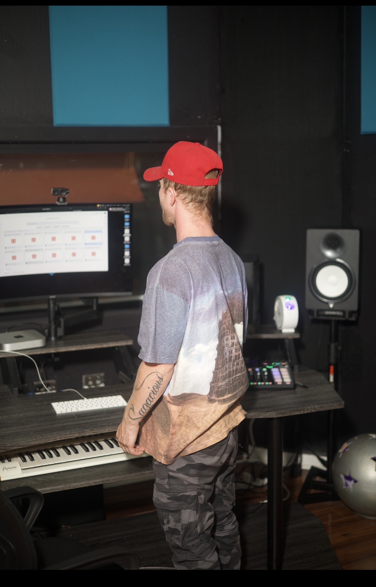 Person wearing a red cap and gray shirt standing in a music studio with a keyboard and computer screen.