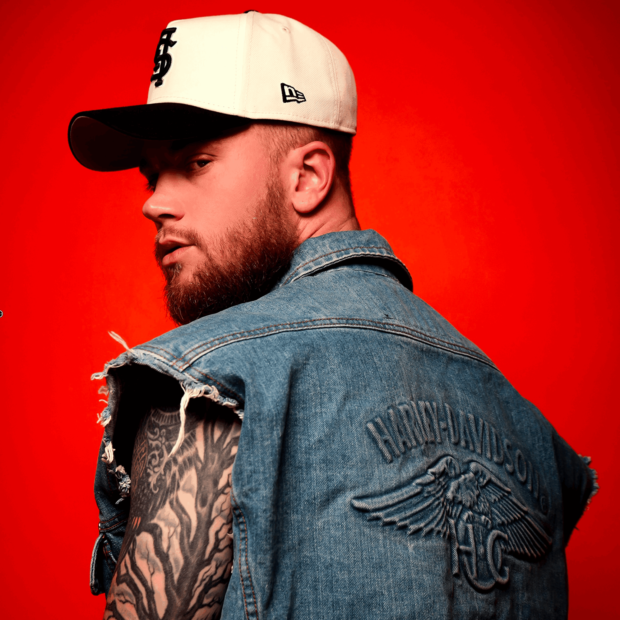 Man with beard wearing a white cap, denim vest, and tattooed arm against red background.