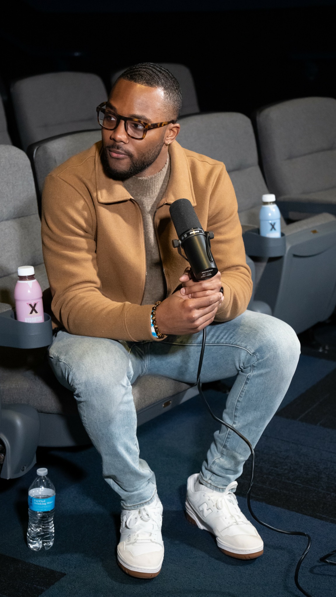 Man with glasses and beard sitting in a theater, holding a microphone, surrounded by water bottles and cups.
