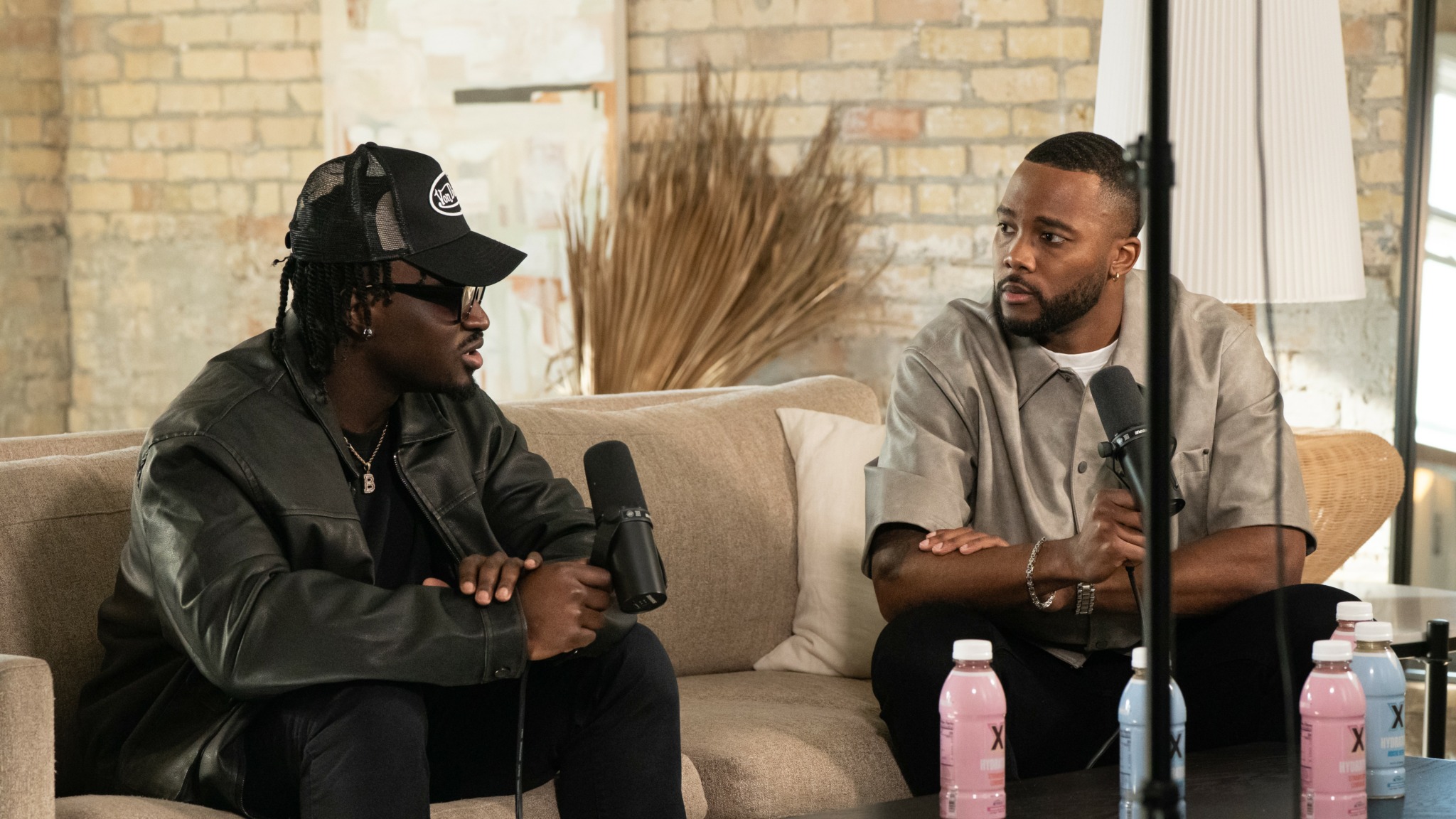 Two men sit on a beige sofa, holding microphones, engaged in conversation indoors with a brick wall and window behind them.