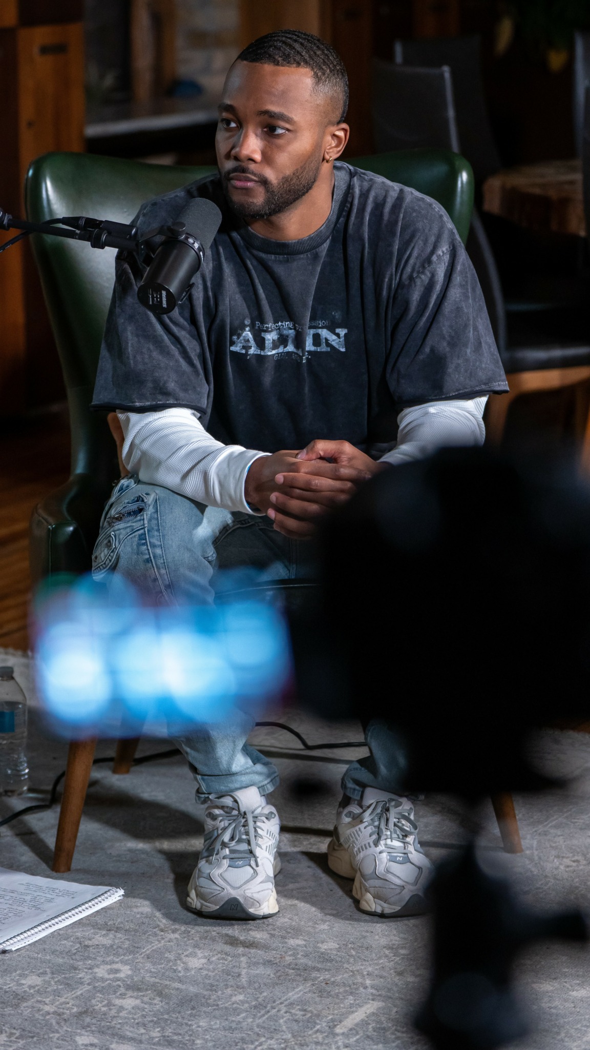 Man with short hair and beard sitting at a table with a microphone, wearing a dark T-shirt and jeans.