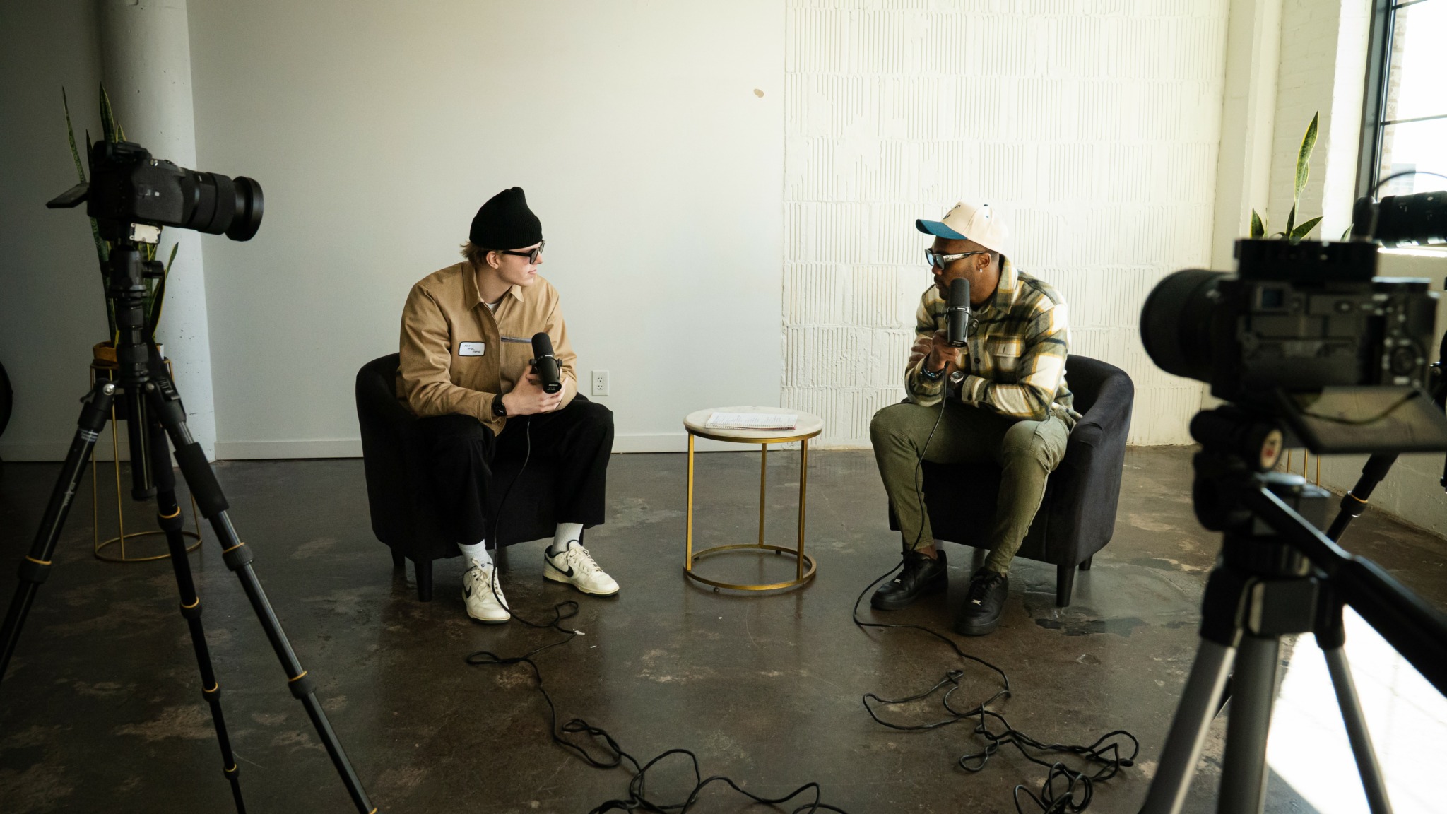Two people sitting in chairs with cameras and equipment around them, in a room with white walls and a window.
