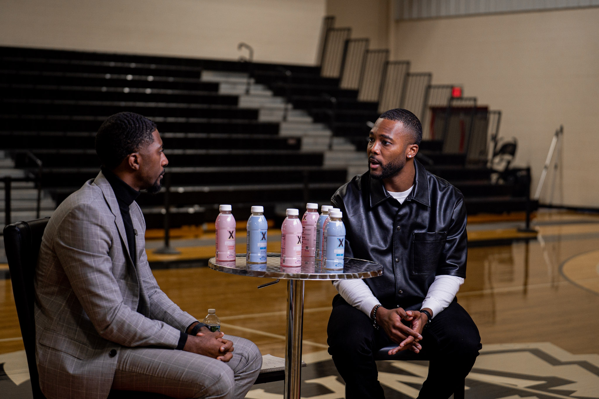 Two men sitting at a table in a gymnasium, engaged in conversation, with water bottles on the table.
