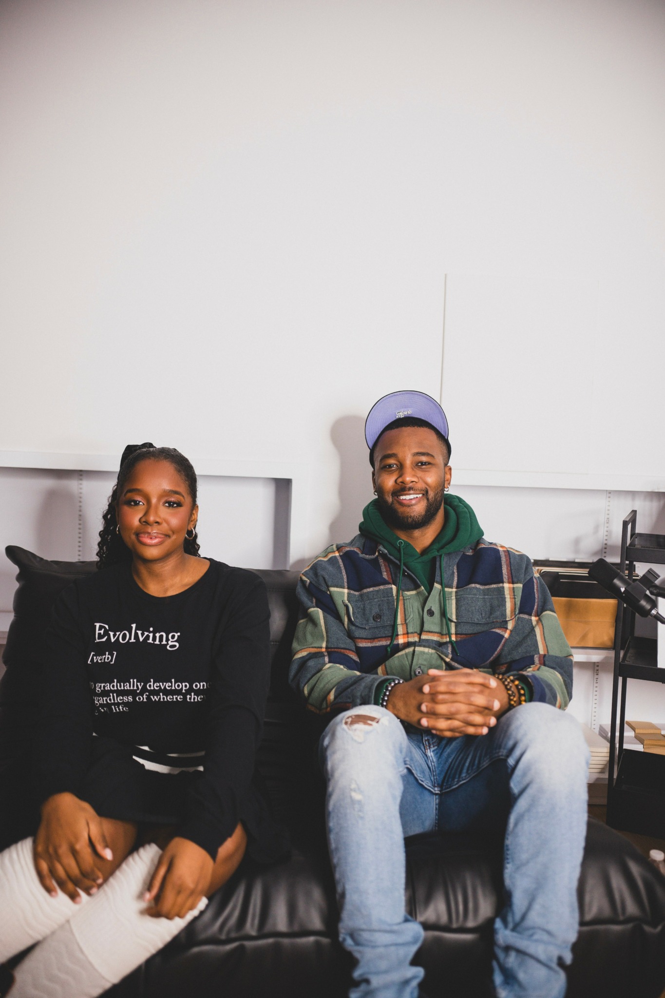 Two people sitting on a black leather couch in a room with a white wall behind them, smiling.