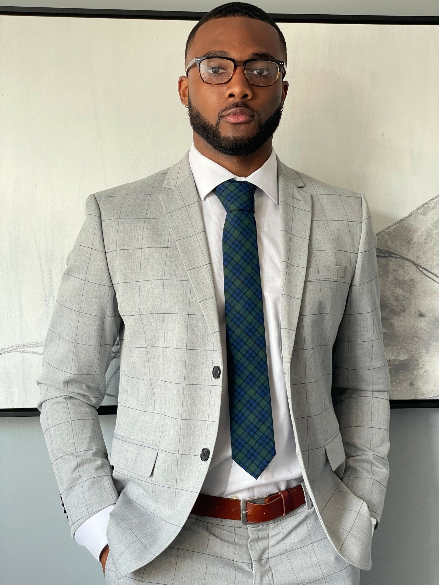 Man in a light gray checked suit, white shirt, and blue plaid tie, standing indoors with a neutral background.
