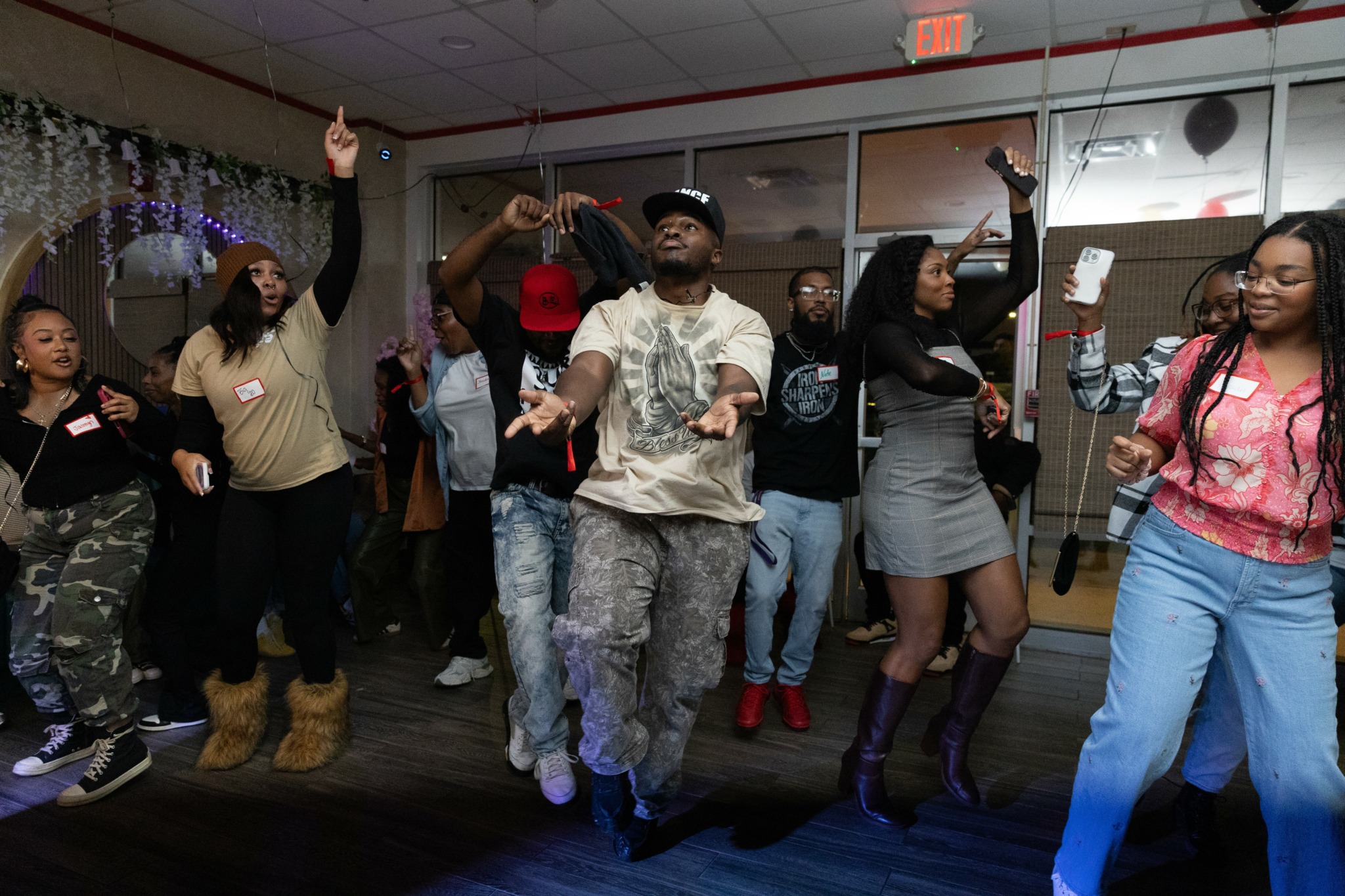 Group of people dancing indoors, some with arms raised, in casual clothing, with a mirror and decorations in the background.