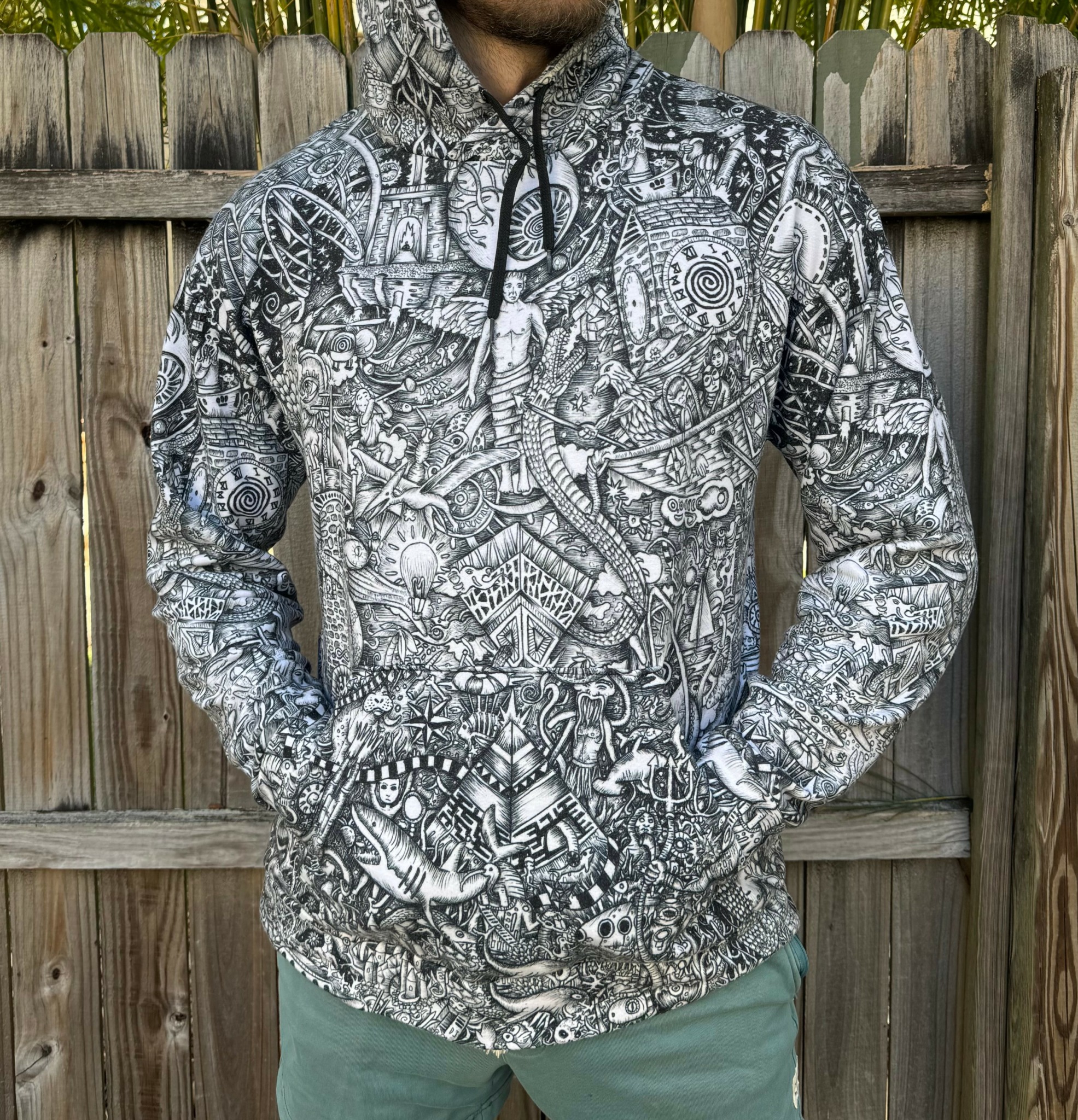 Person wearing a patterned hoodie with hands in pockets, standing in front of a wooden fence.
