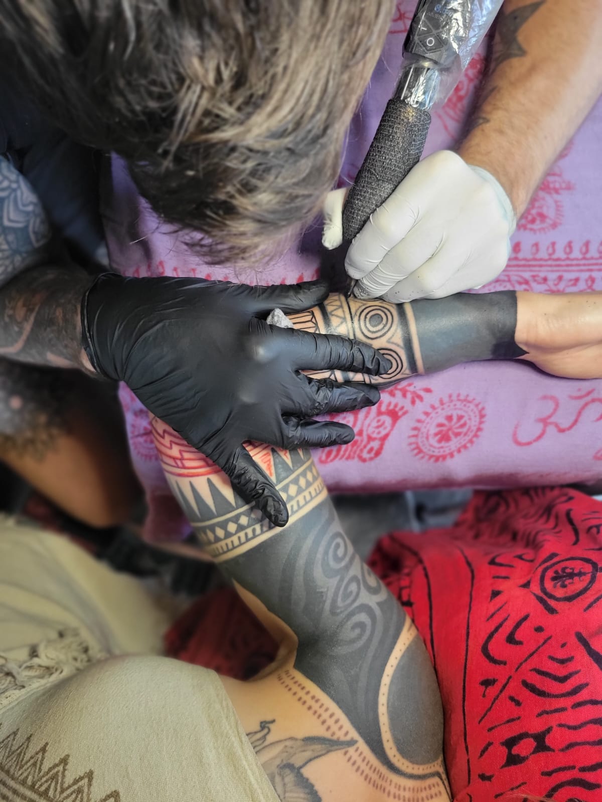 Tattoo artist in black gloves working on a person's arm with intricate black ink designs.