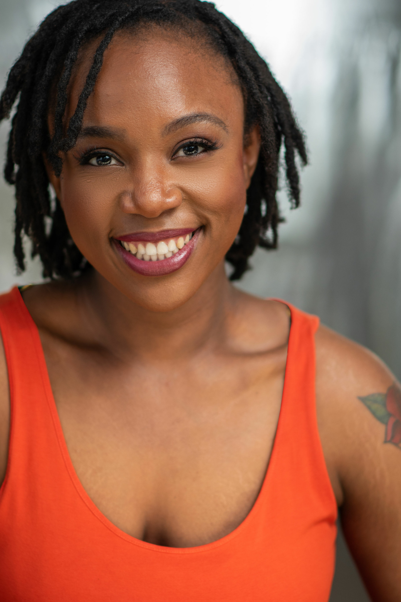 Smiling woman with dreadlocks wearing an orange tank top, outdoors with blurred background.