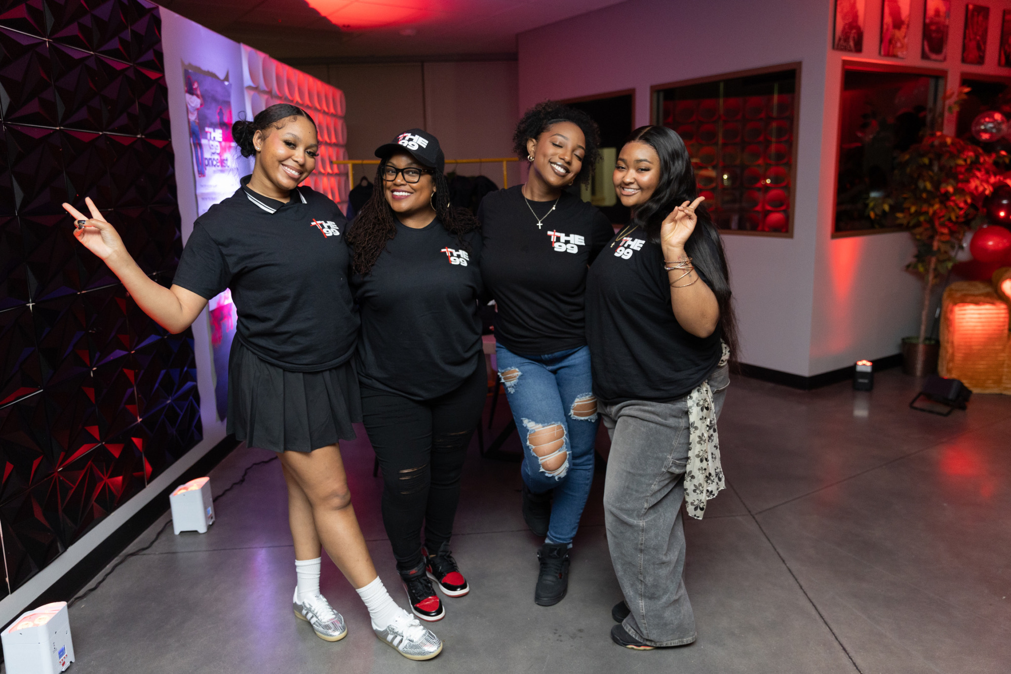 Four women standing together indoors, smiling, with one making a peace sign, in a room with red lighting and decorations.