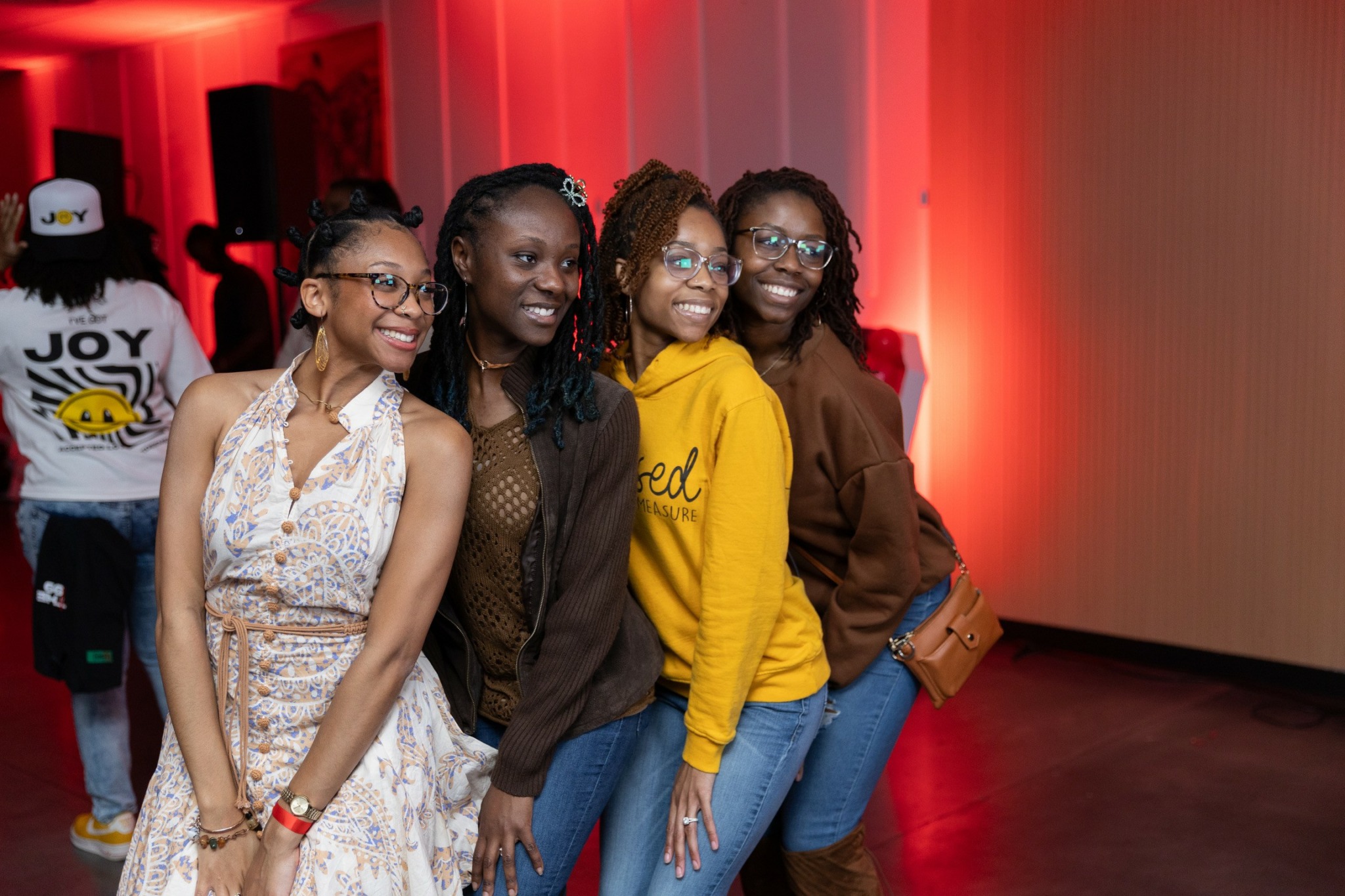 Four women standing together, smiling, in a room with red lighting and a wooden floor.