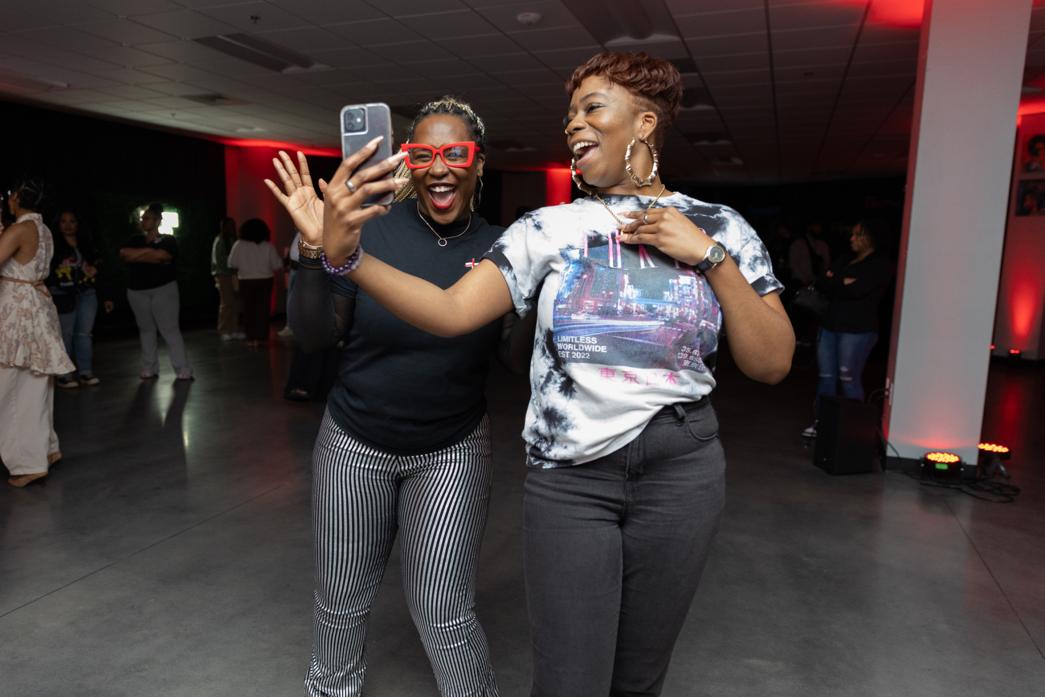 Two women smiling and taking a selfie together at an indoor event, with other people in the background.