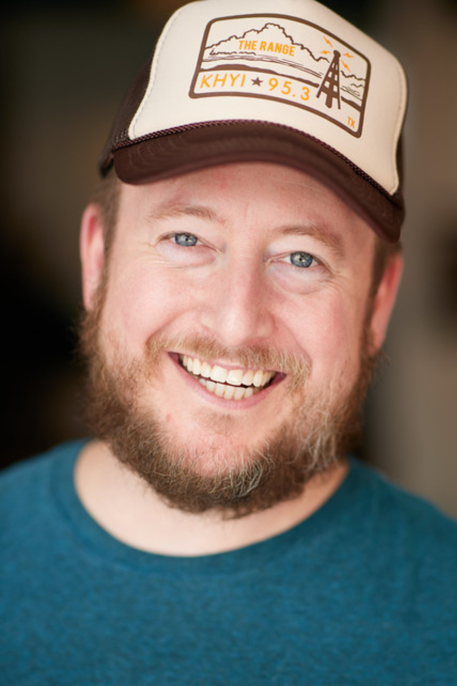 Smiling man with beard wearing a cap with a bridge illustration and text, and a blue shirt.