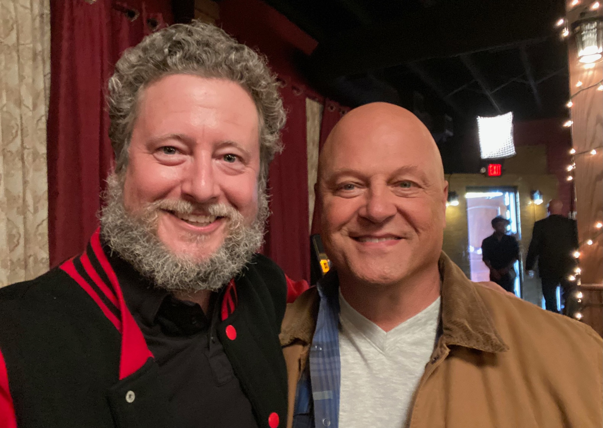 Two men smiling indoors, one with curly gray hair and beard, the other bald, in casual clothing.