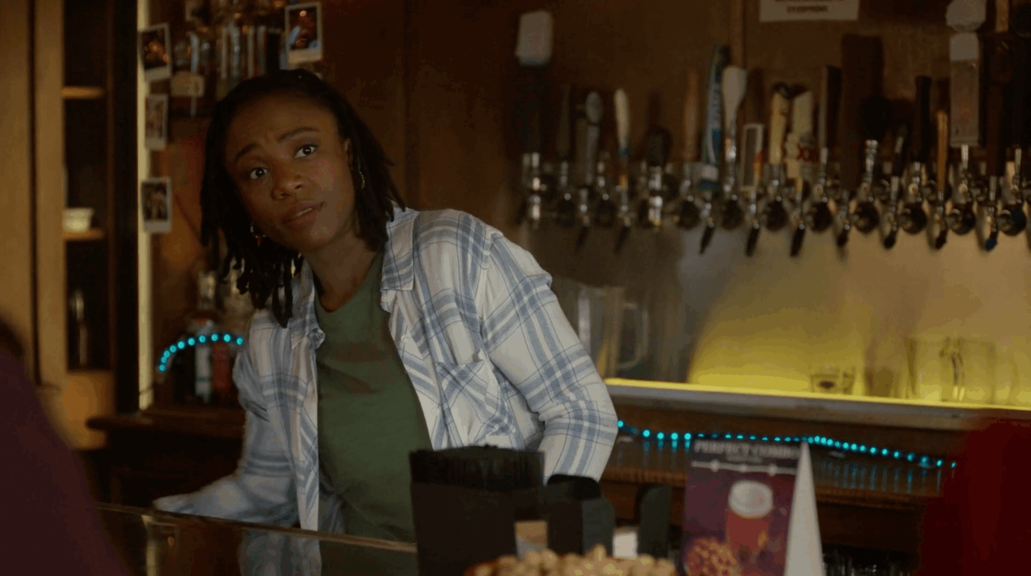 Woman with dreadlocks sitting at a bar counter, wearing a plaid shirt and green top, looking to the side.