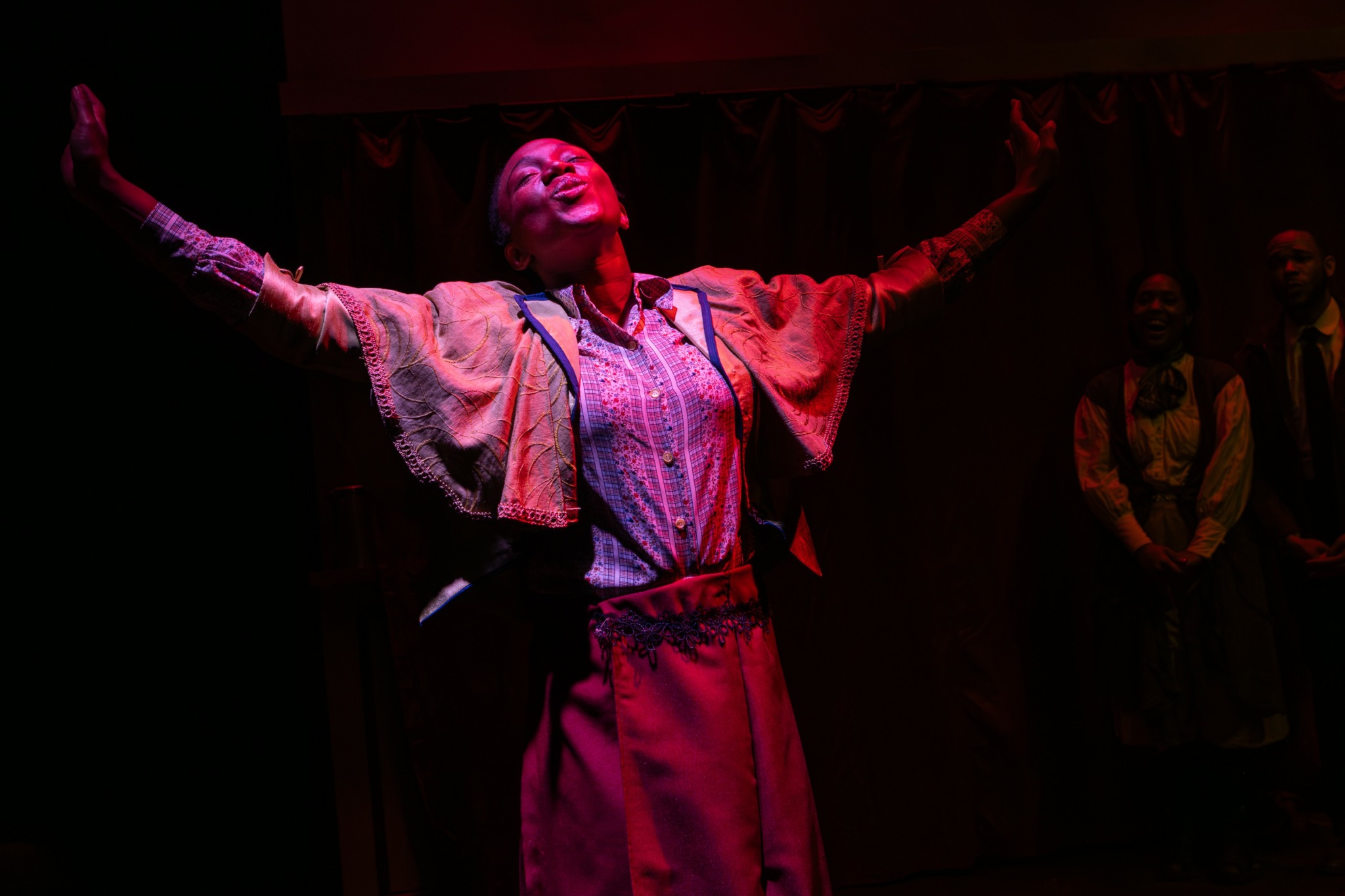 Performer on stage with arms raised, wearing a patterned jacket and skirt, with dark background and other people in the background.