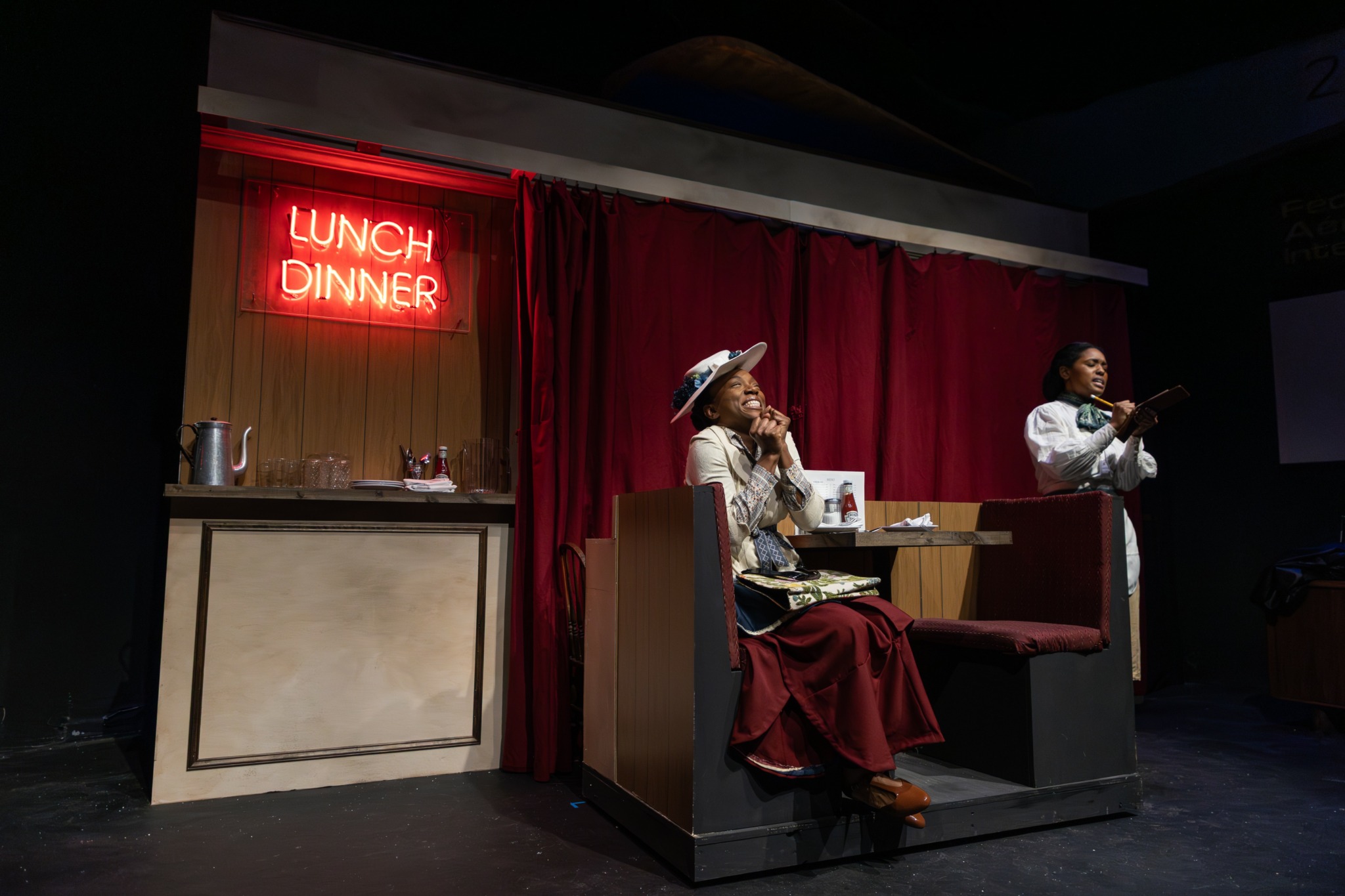 Two women perform on stage with a red curtain backdrop, one singing and the other playing guitar, near a lunch and dinner sign.