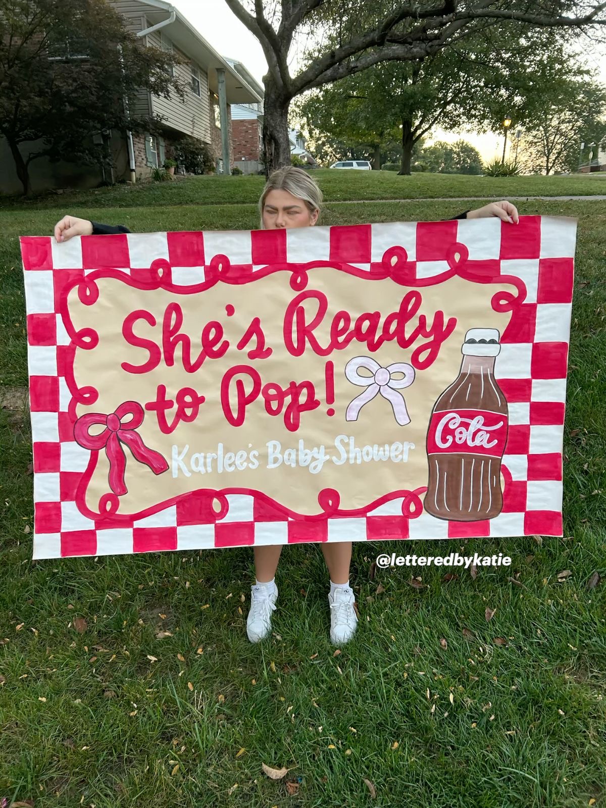 Person holding a large colorful sign with a Coca-Cola bottle illustration and text, outdoors on grass with trees in background.