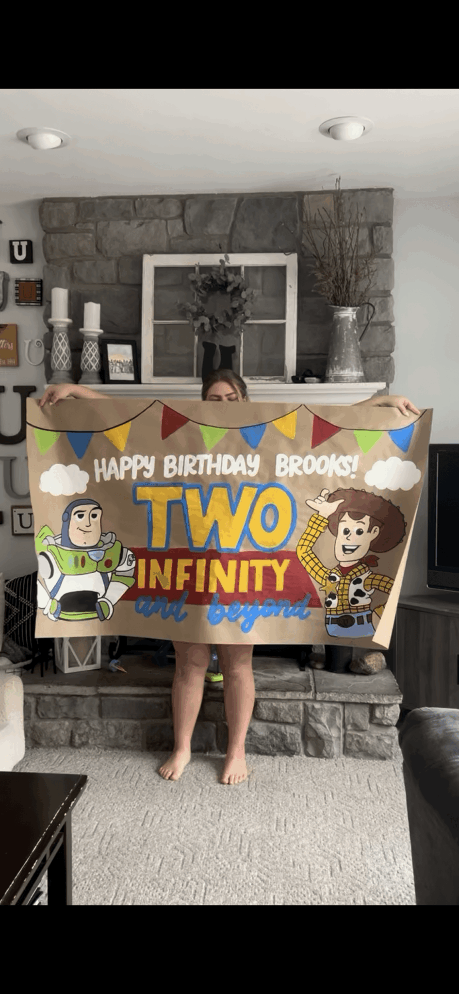Child holding a Two Infinity birthday banner in a living room with stone fireplace and decorations.