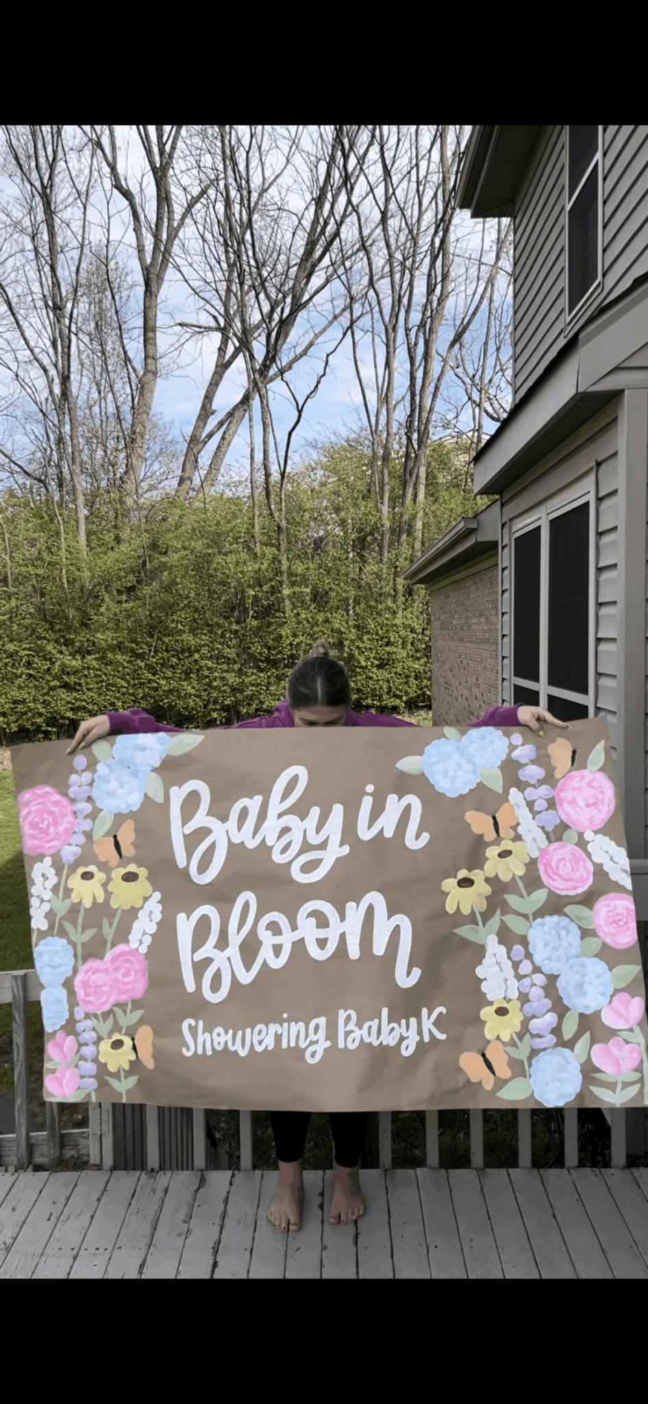 Person holding a colorful sign with flowers and text, standing outdoors near a house and trees.