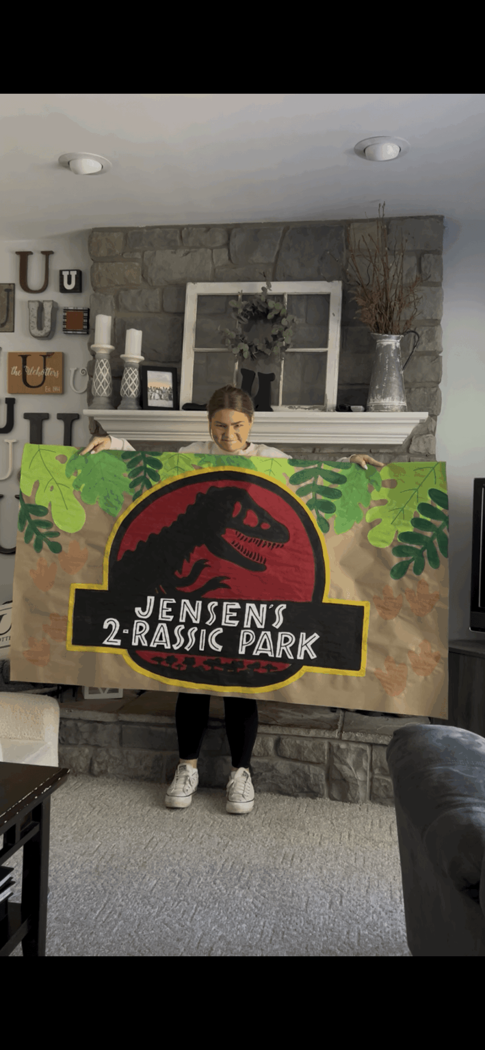 Child holding a large Jurassic Park-themed sign indoors with stone wall and decorative items behind.