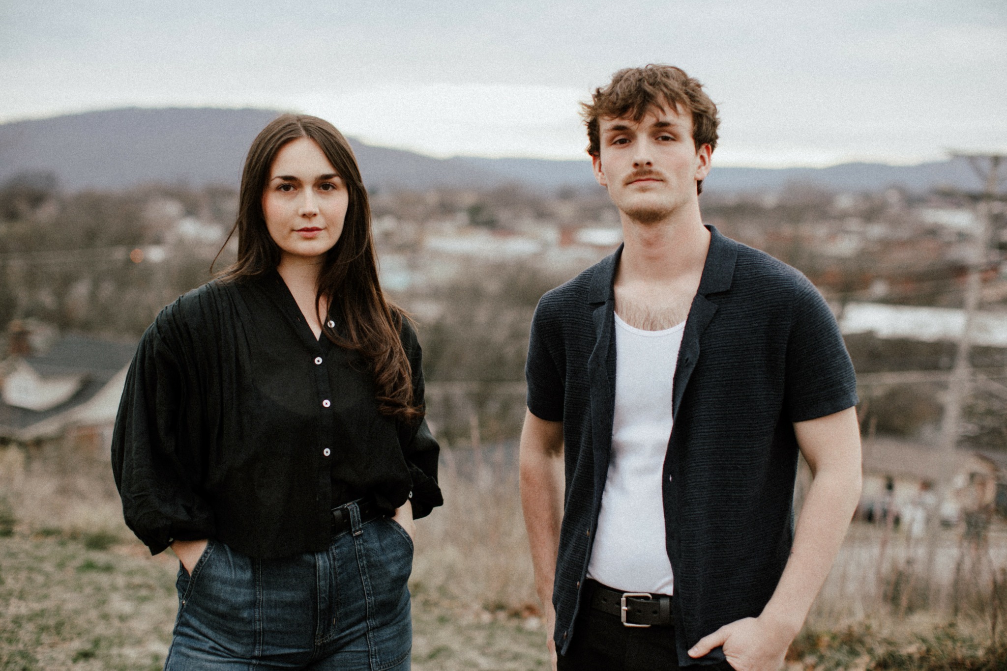 A man and woman stand outdoors with a landscape background, both facing forward.