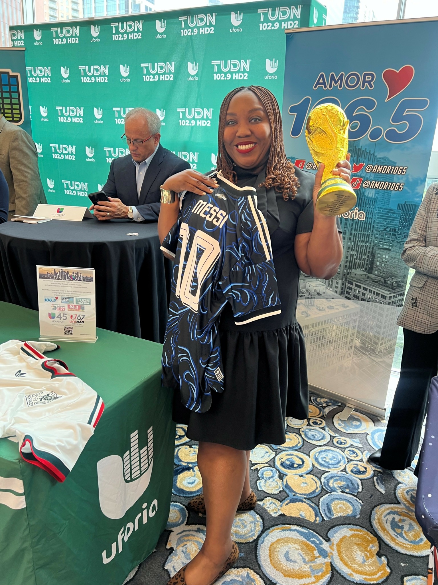 Woman smiling holding a yellow glove and sports jersey, standing in front of a blue promotional backdrop, with a table and people nearby.