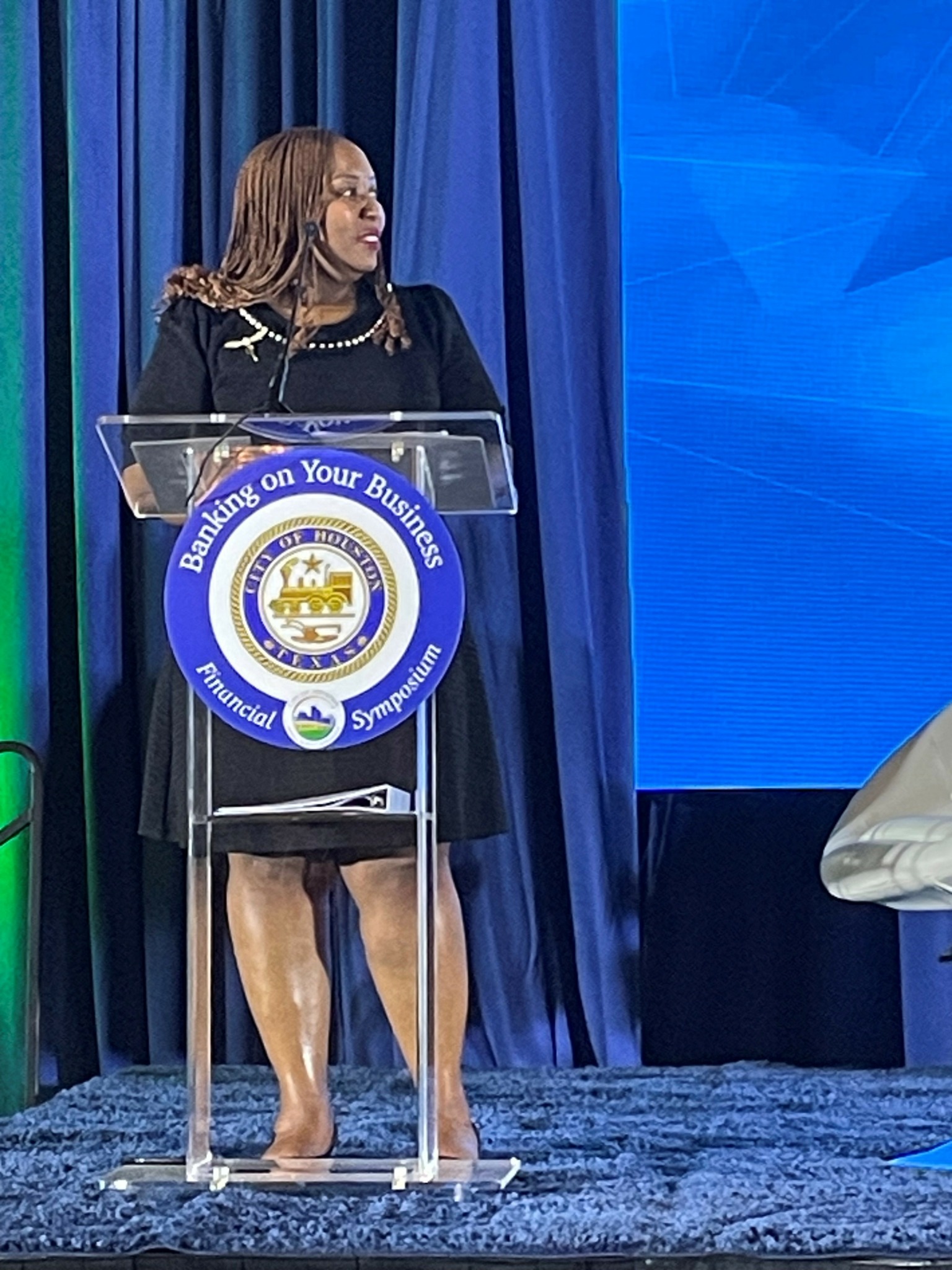 Woman standing at a clear podium with a blue emblem, speaking at an event with blue curtains and a large blue screen behind.