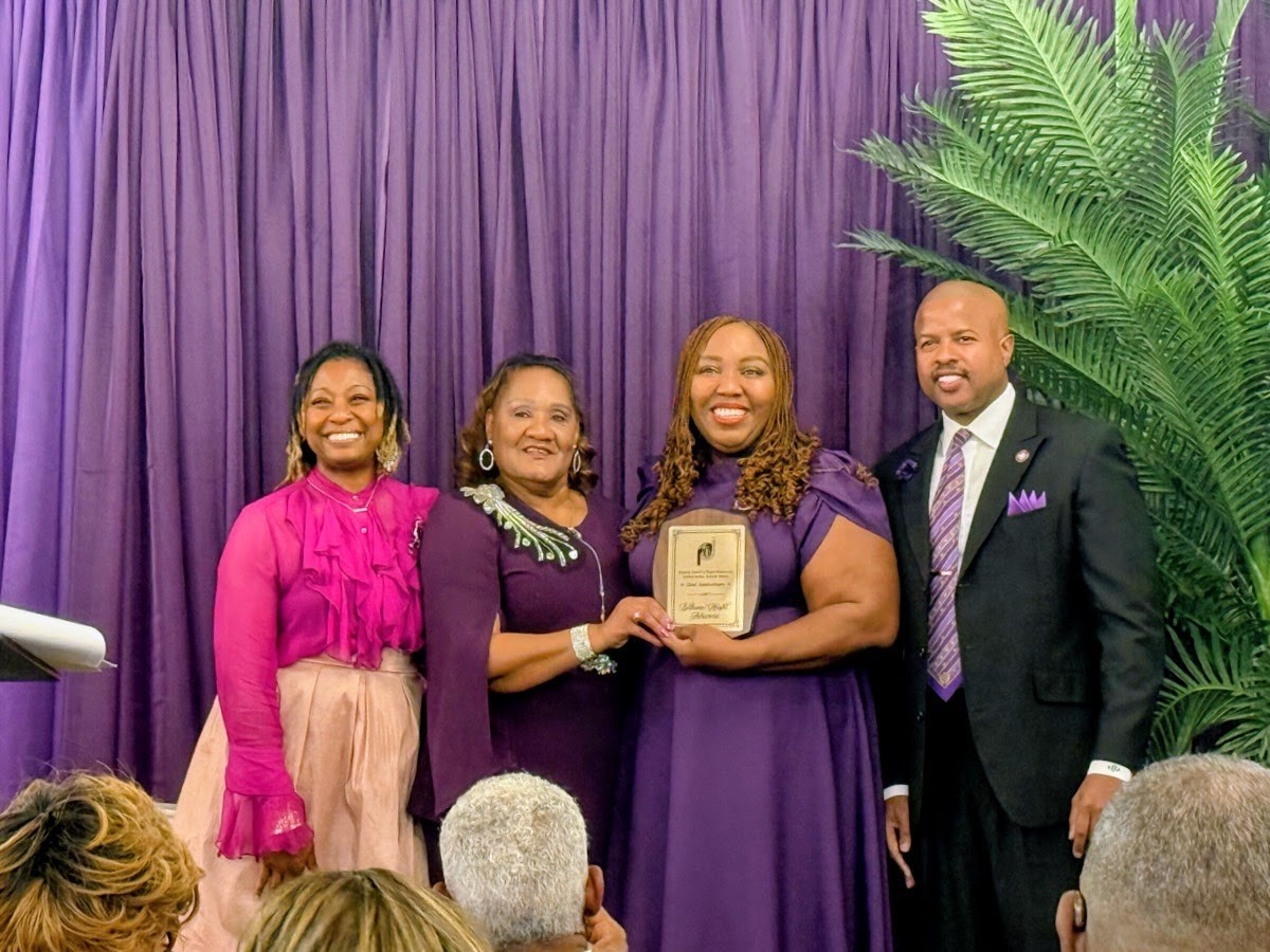 Four people stand in front of purple curtains, smiling, with a large green plant on the right side.