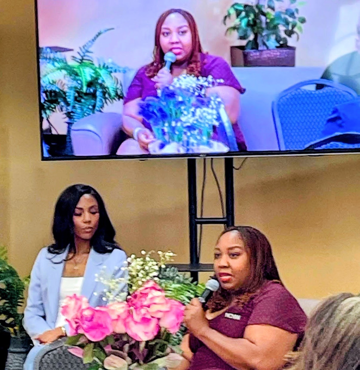 Two women seated, one speaking into a microphone, with a large screen showing her image above them, and flowers in front.