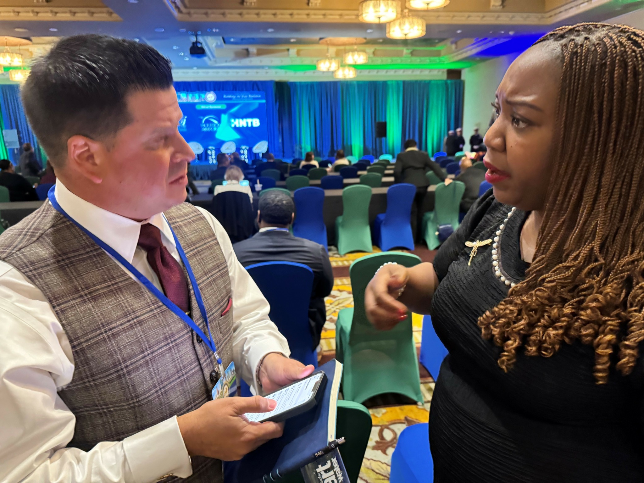 Two people engaged in conversation at a conference with a stage and audience in the background.