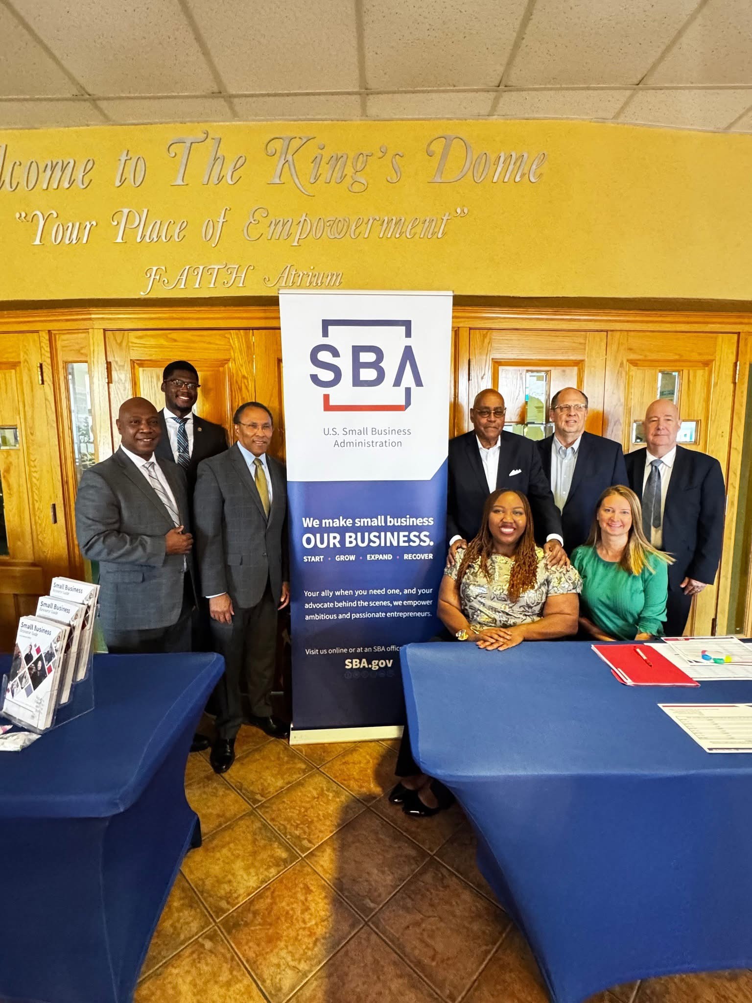 Group of eight people, six men and two women, standing and sitting behind a table with SBA banner, in a room with yellow wall and wooden paneling.
