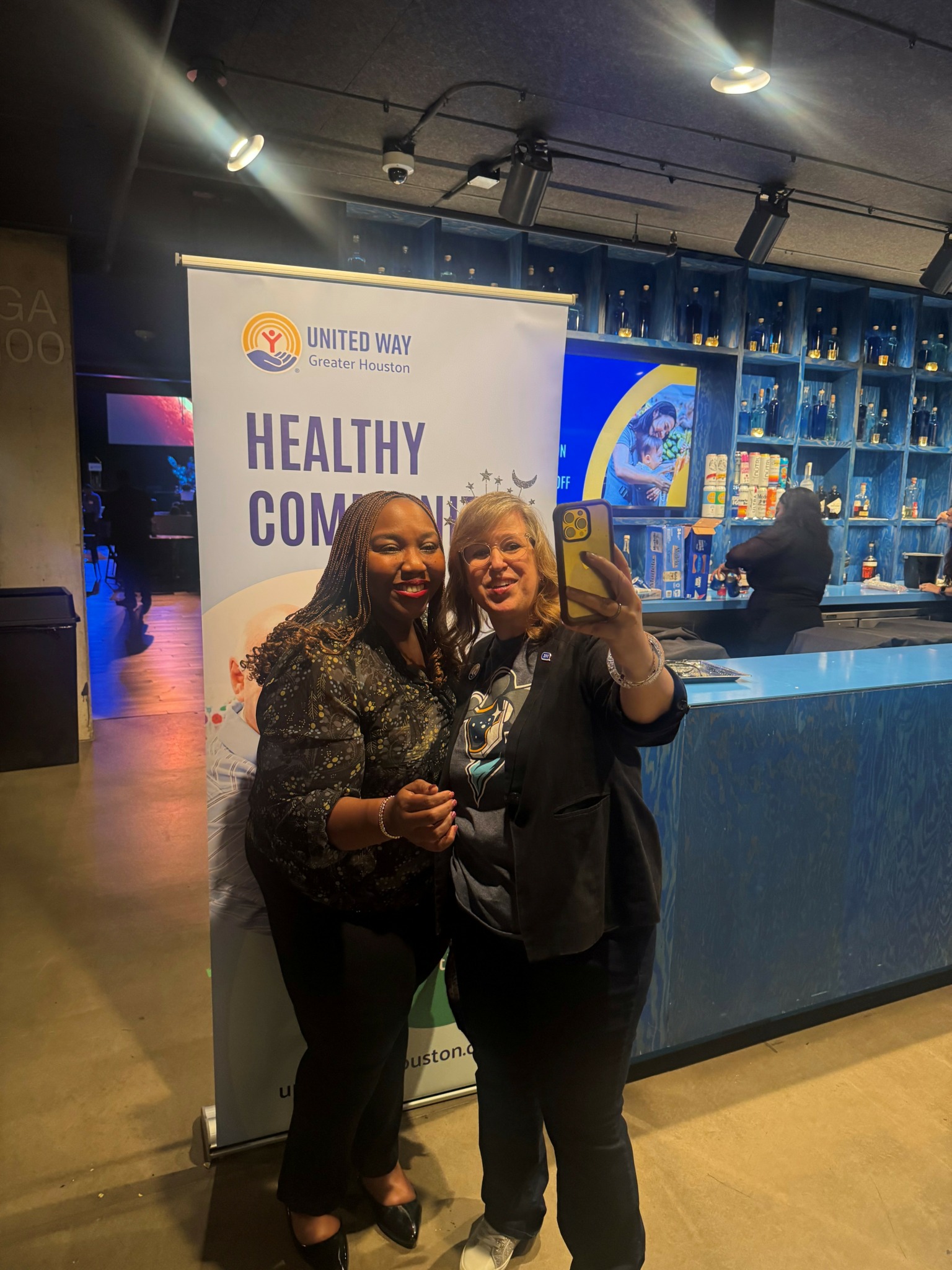 Two women taking a selfie in front of a banner at an indoor event, smiling and standing close together.