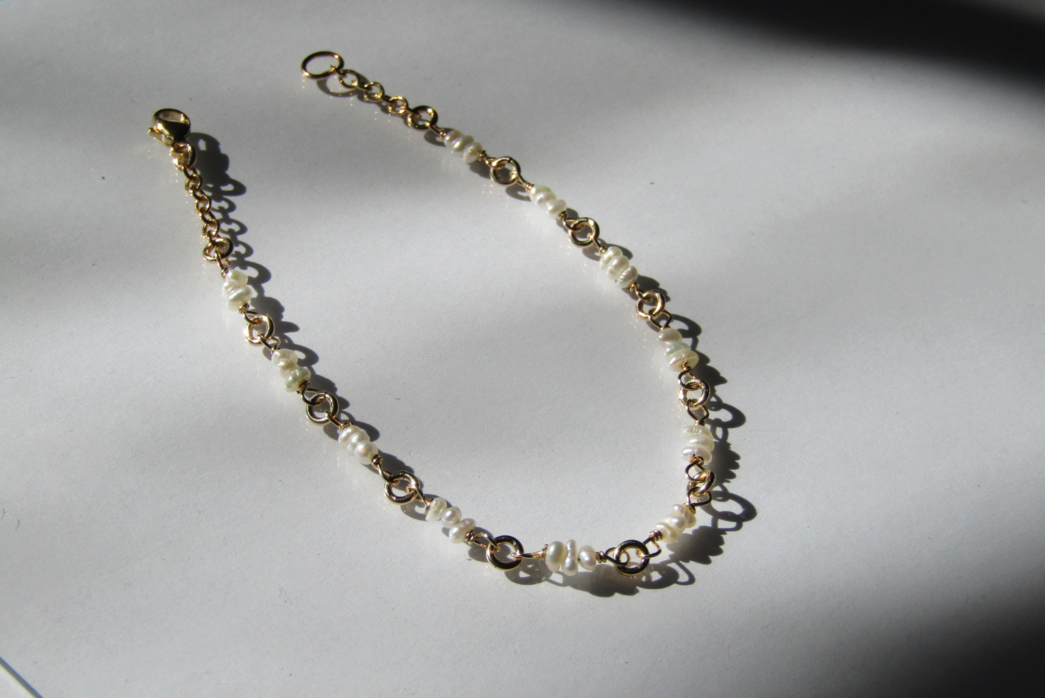 Gold chain necklace with small beads and charms, laid on a white surface with shadows.