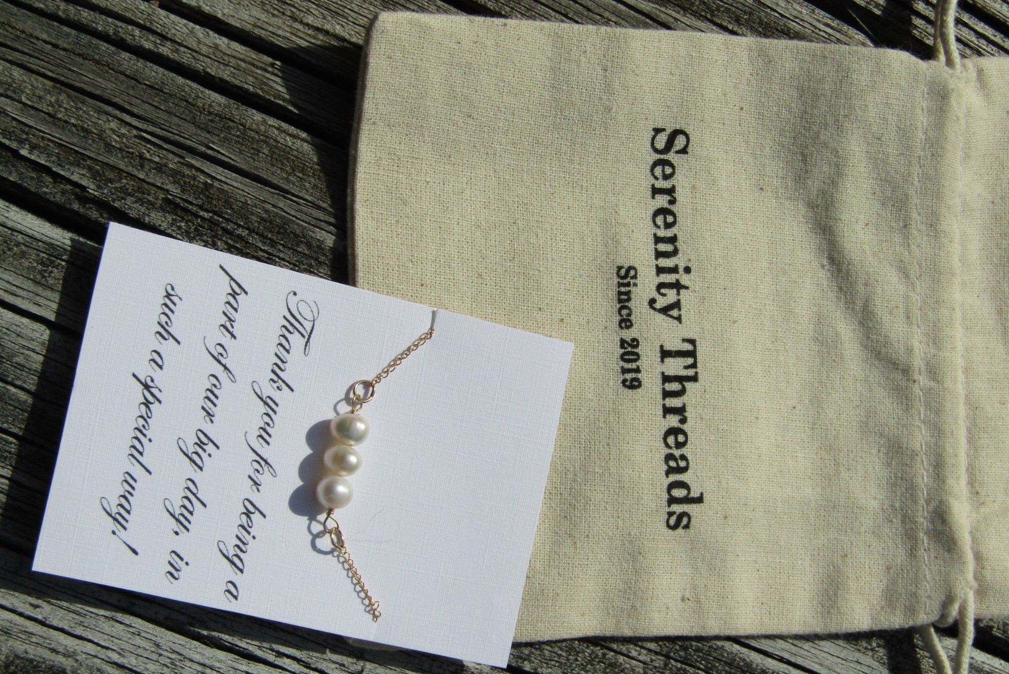 Pearl necklace on a card with handwritten message, beige cloth bag with black text, wooden surface background.