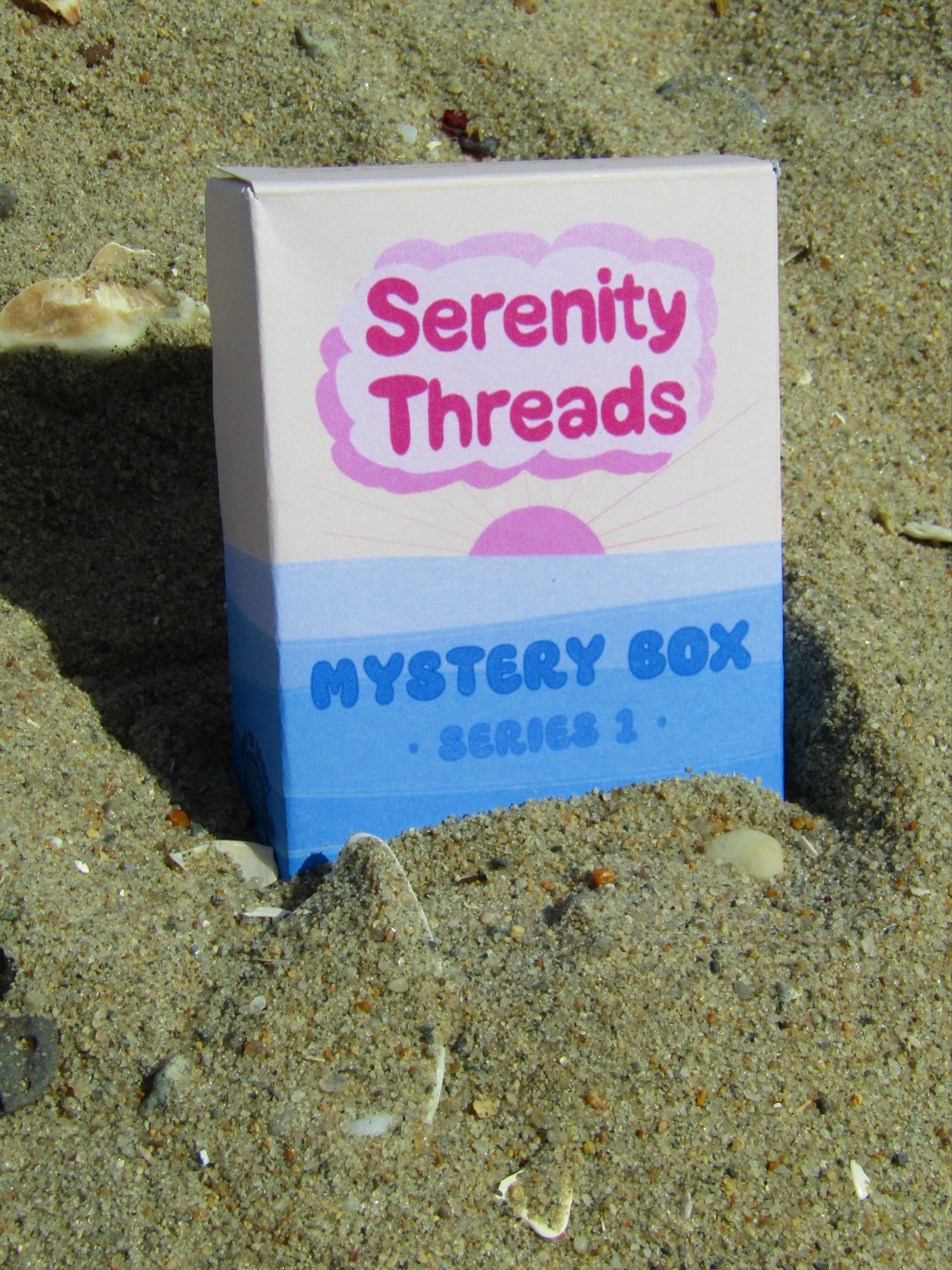 Box labeled 'Serenity Threads Mystery Box Series 1' partially buried in sand on a beach.
