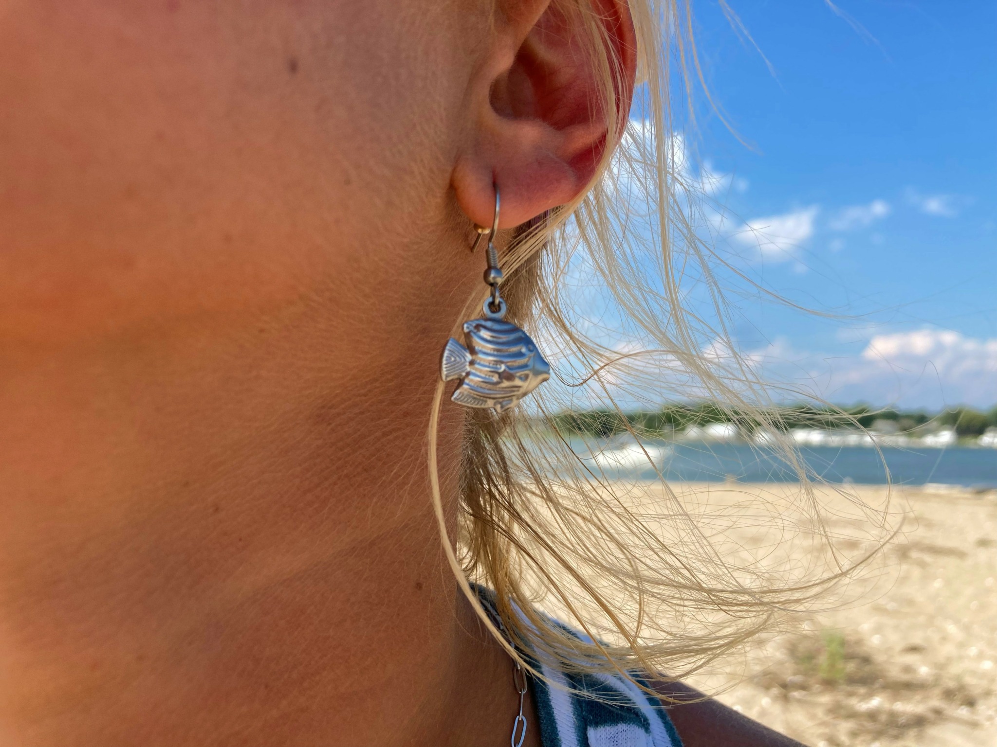 Close-up of a person's ear with a fish-shaped earring, outdoors on a beach with blue sky and sand.