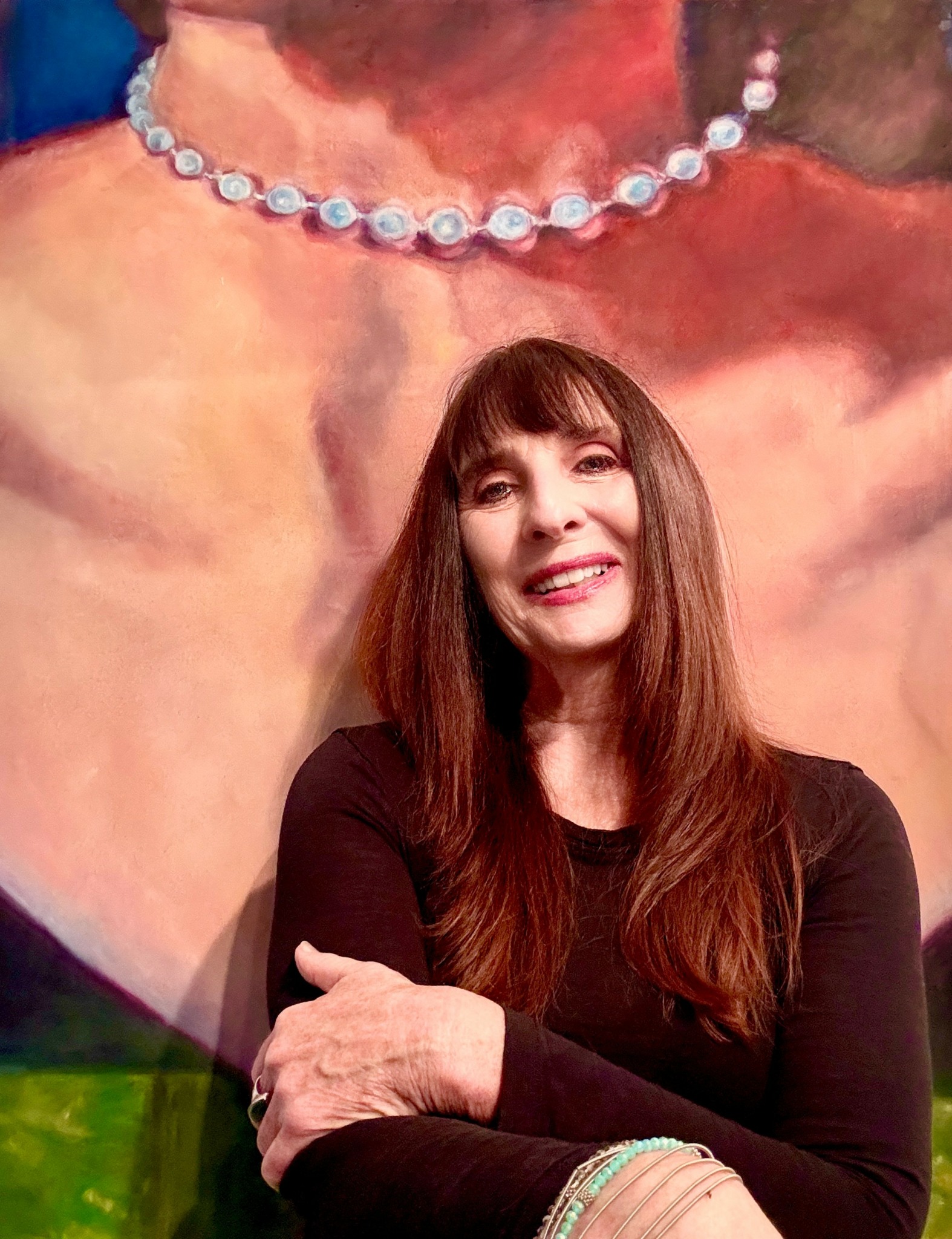Woman with long brown hair smiling, wearing a black top, in front of a large painted portrait with a pearl necklace.
