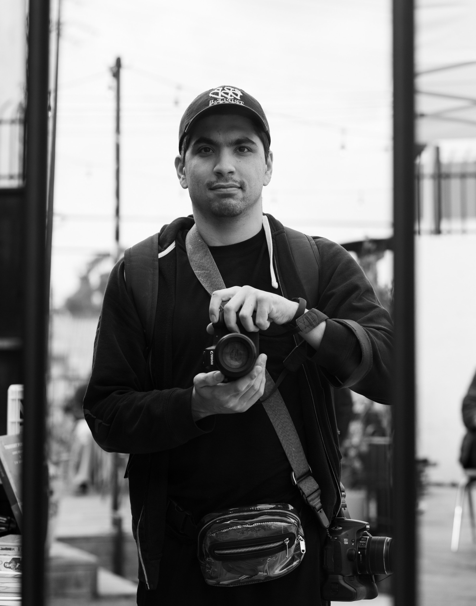 Young man holding a camera, wearing a cap and backpack, standing outdoors with a blurred background.