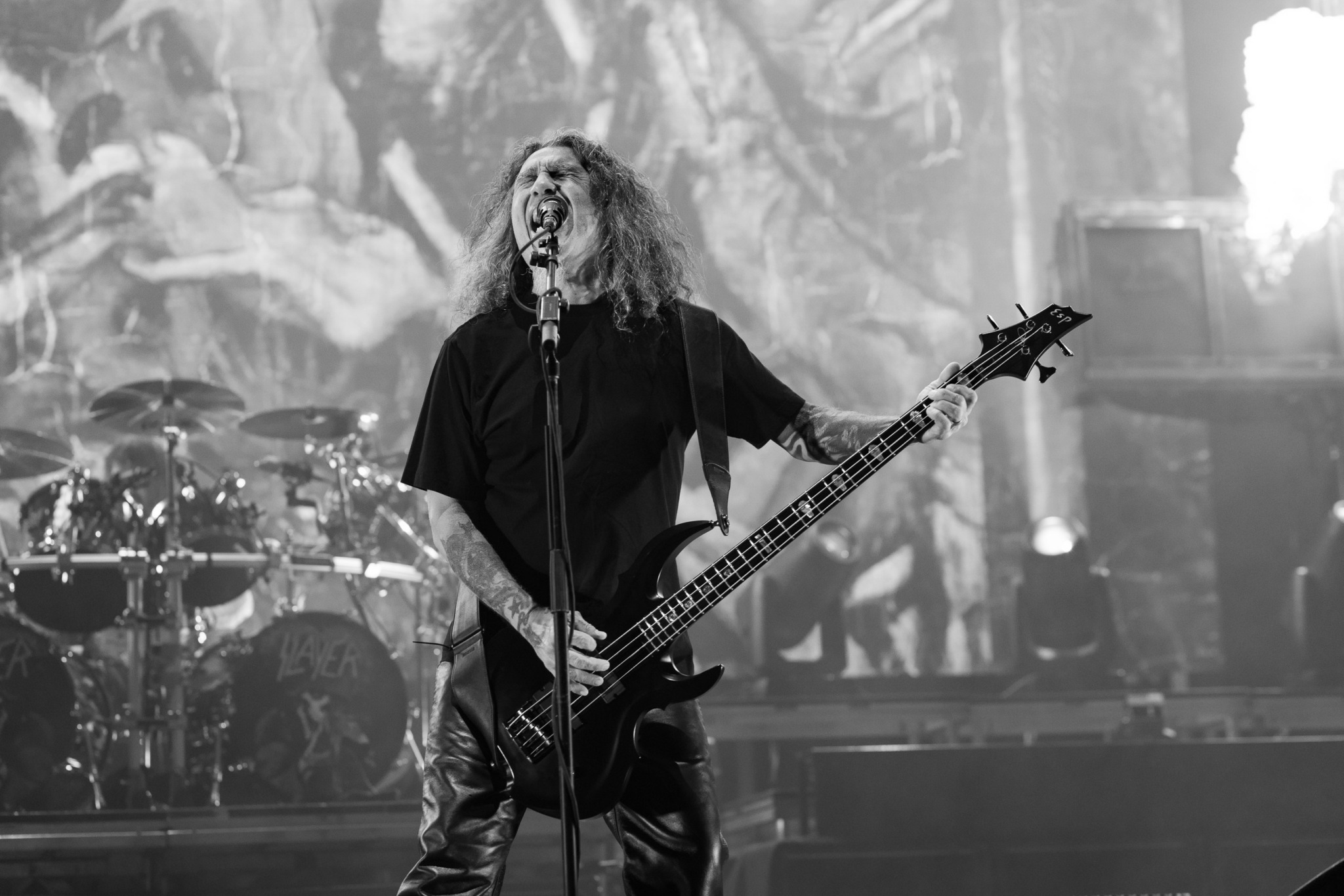 Musician with long curly hair playing bass guitar on stage, with drums in background, in black and white.