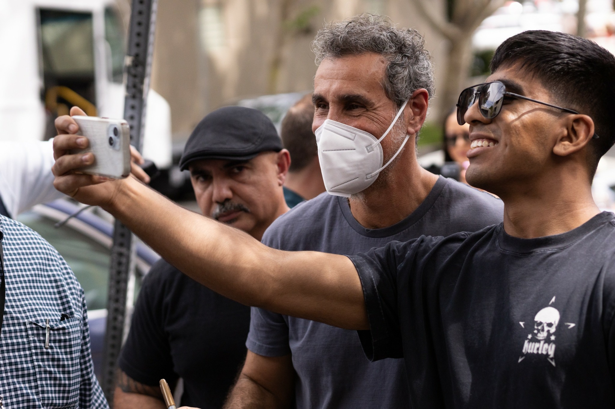 Group of people outdoors, one person taking a selfie, wearing sunglasses and a face mask, others smiling and looking at the camera.