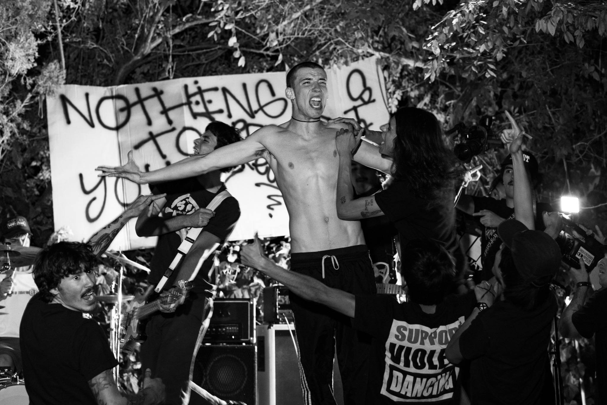 Group of people protesting outdoors with a large sign, some holding hands and raising arms, trees in background.