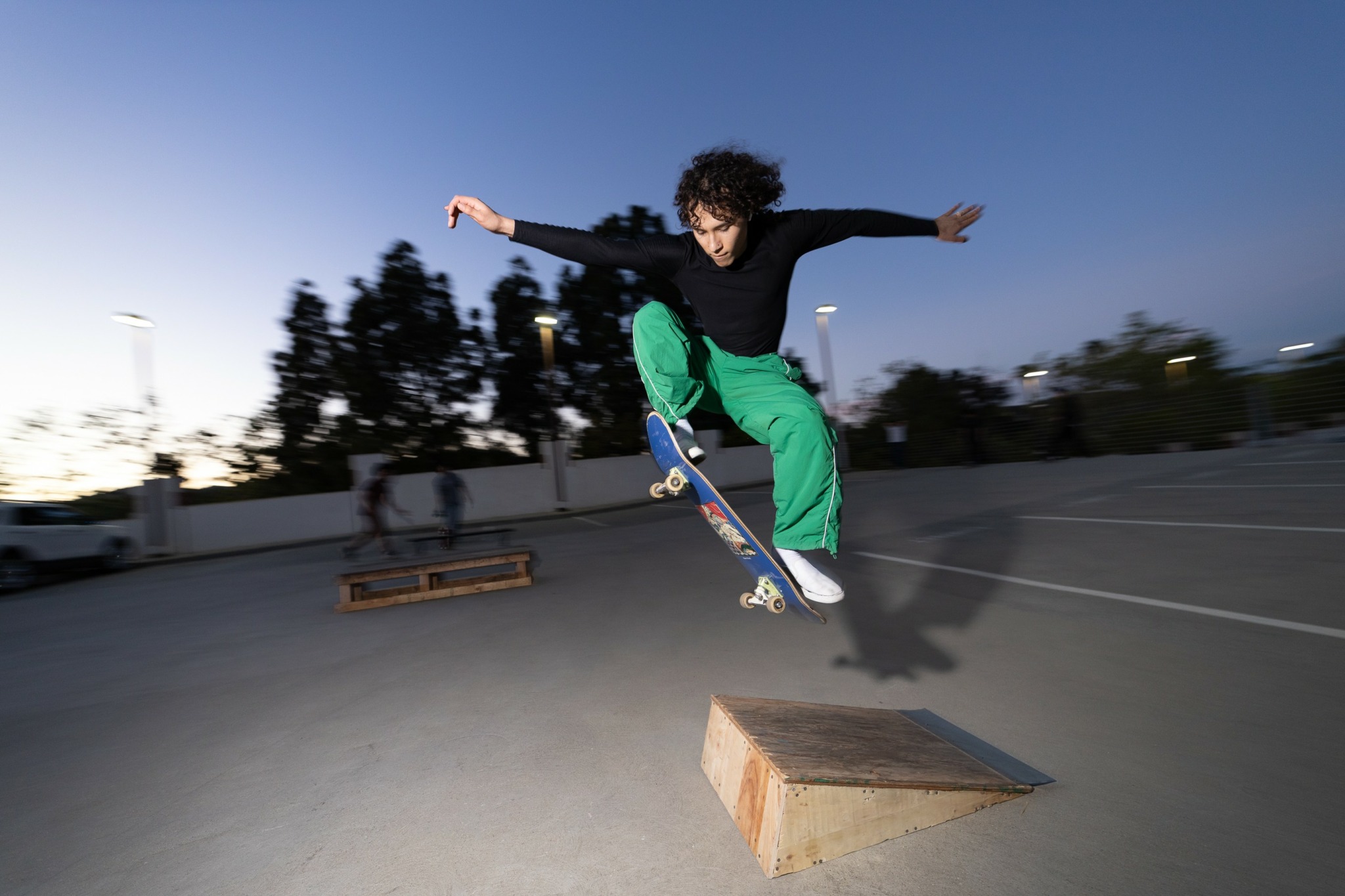 Skateboarder mid-air performing a trick on a ramp outdoors during evening, with trees and sky in background.