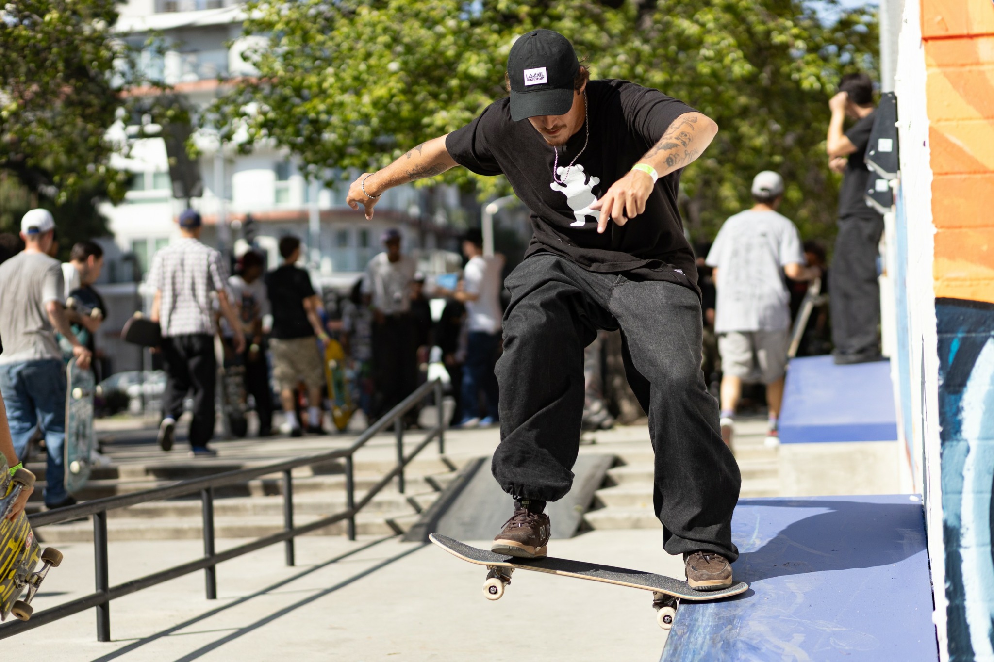 Skateboarder performs a trick on a ledge in an outdoor urban area with people walking in the background.