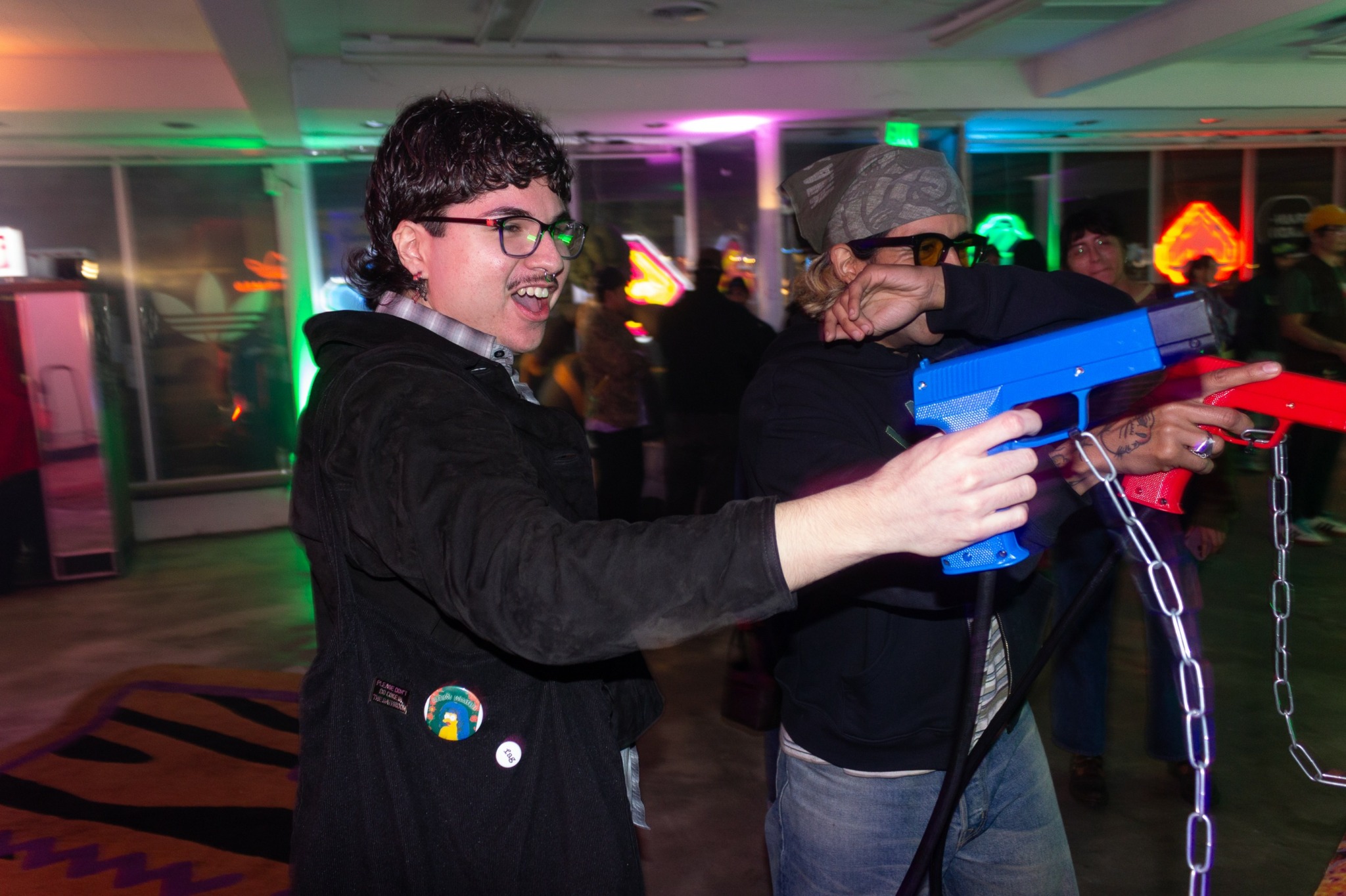 Two people playing laser tag indoors, one smiling and aiming a blue laser gun, colorful lights in background.