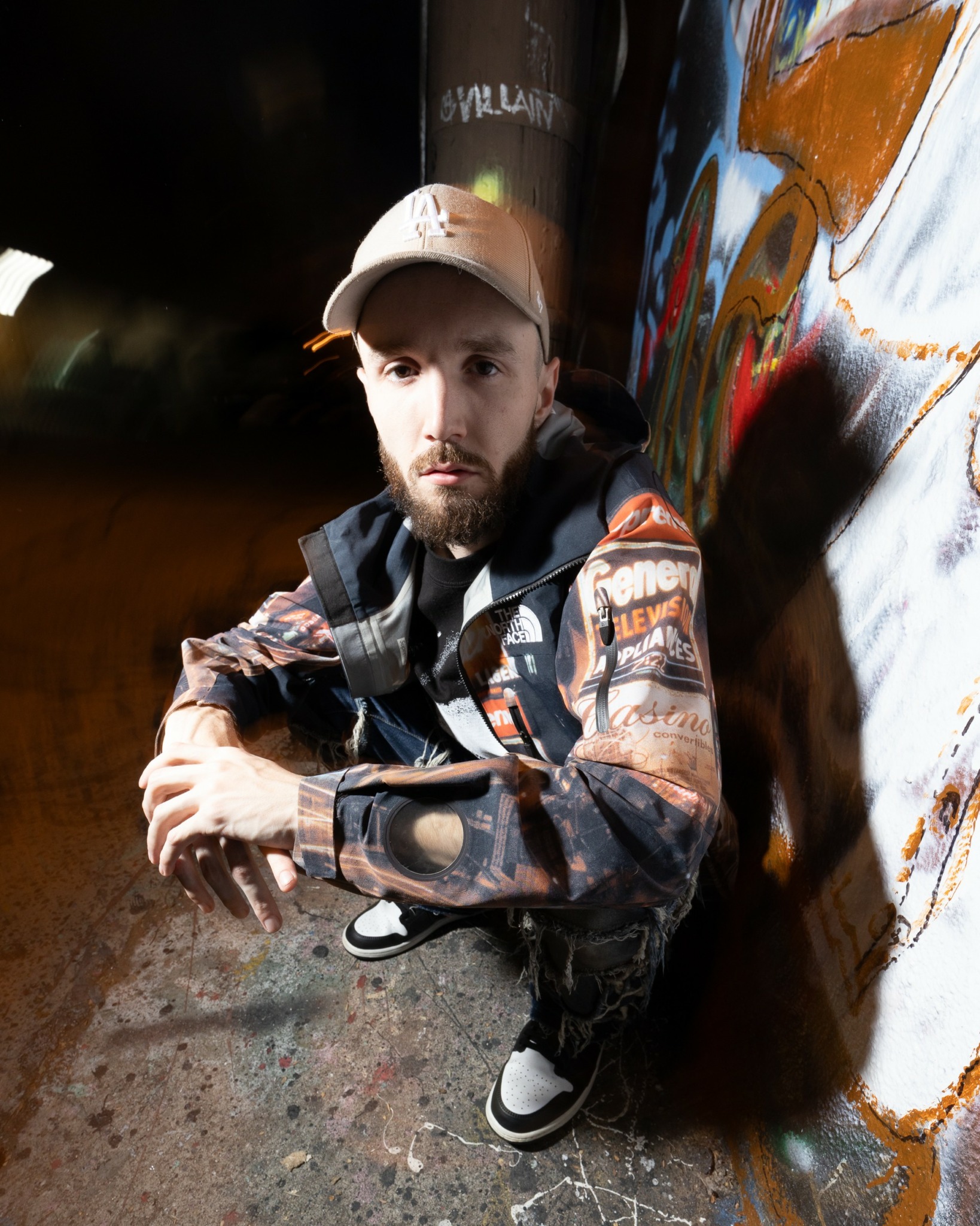 Young man with a beard wearing a cap, jacket, and backpack leaning against a graffiti-covered wall at night.