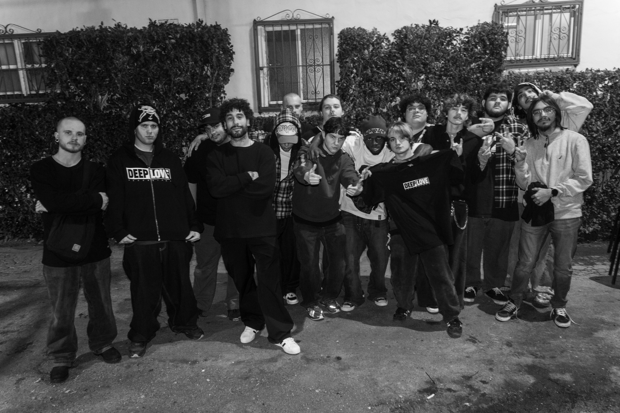 Group of people standing outdoors in front of bushes and windows, some wearing hoodies and jackets.