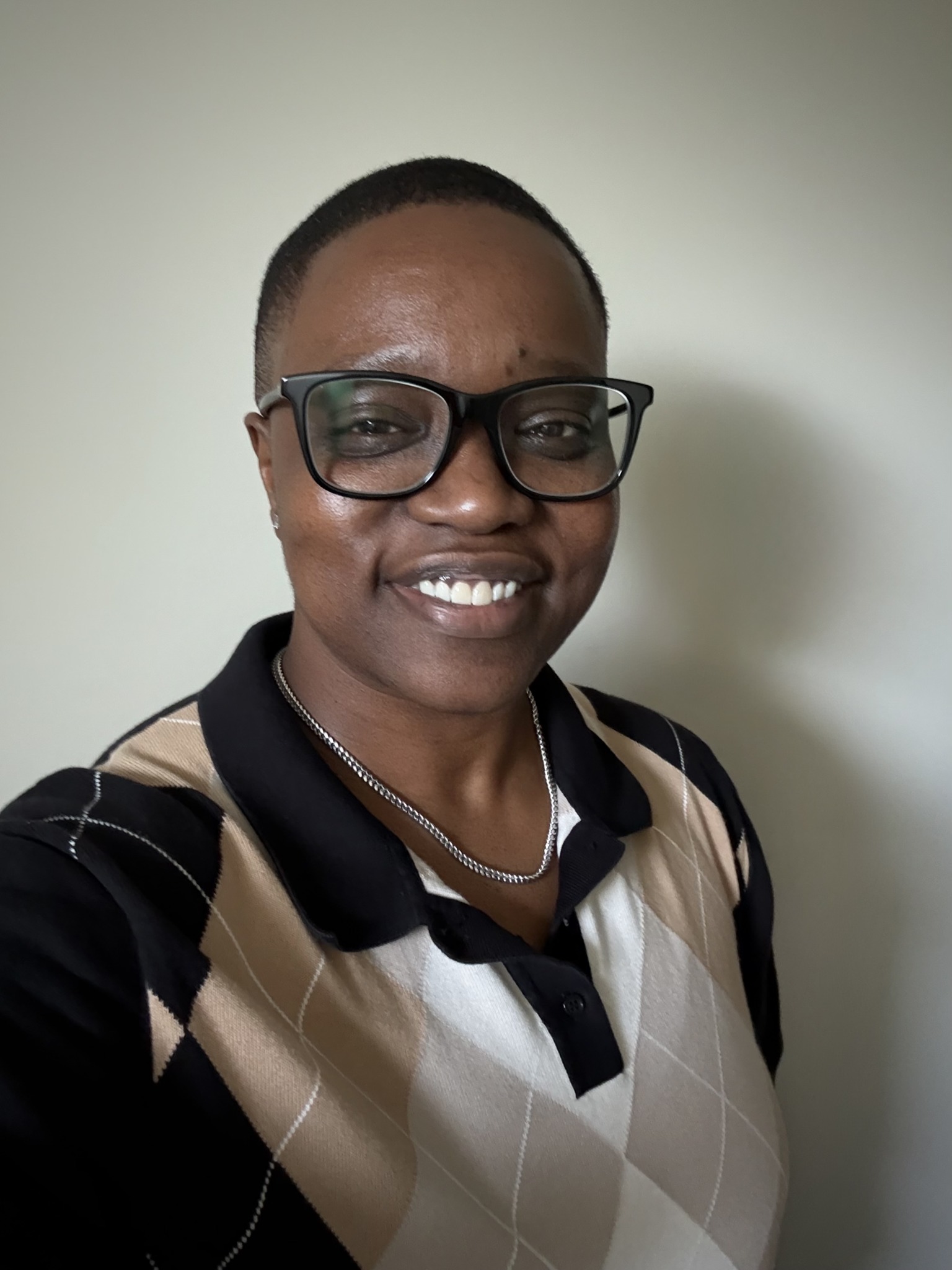 Person with short hair, glasses, smiling, wearing a patterned shirt, standing against a plain background.