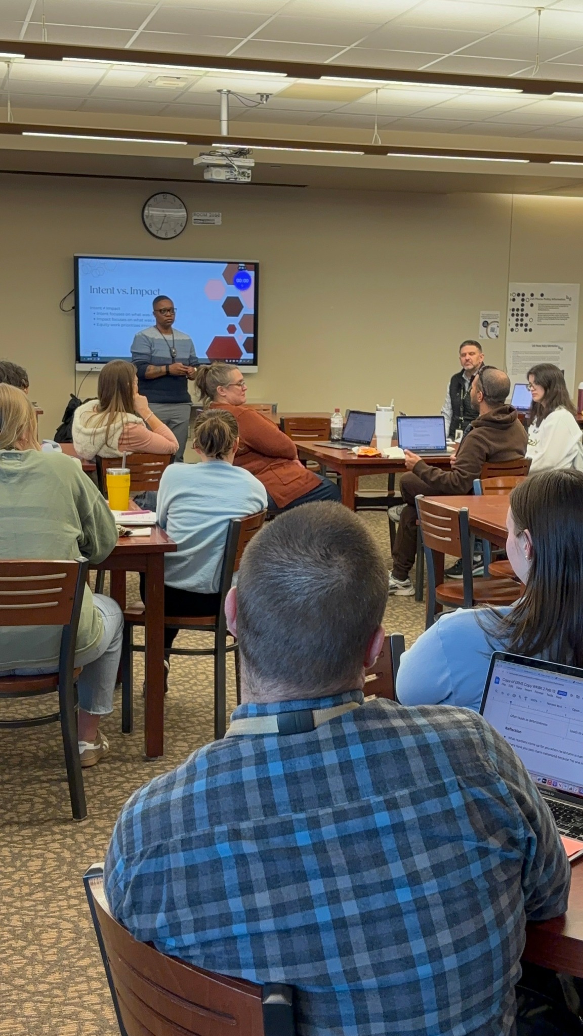 Classroom with people seated at tables, a presenter at the front, and a screen displaying a presentation.