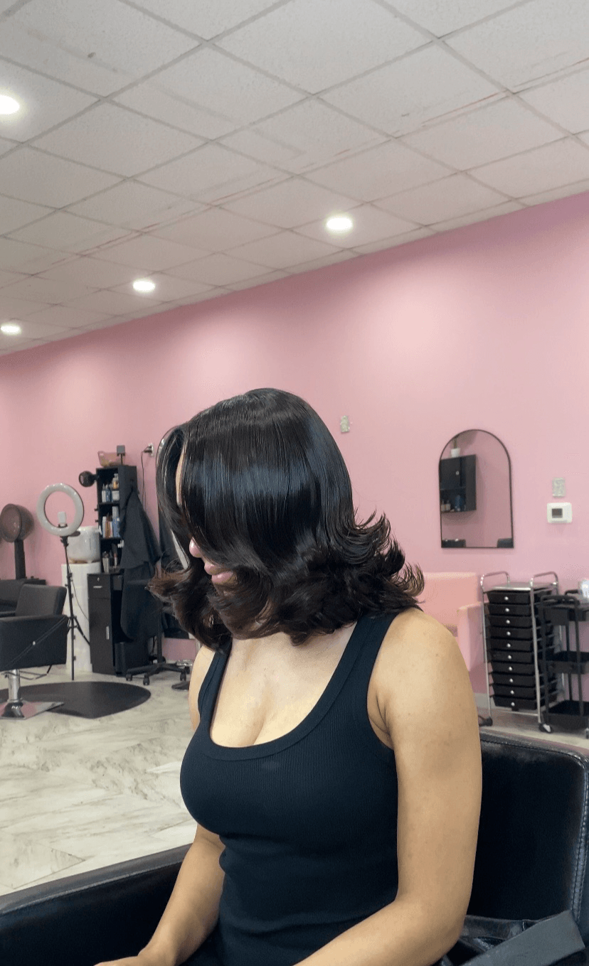 Woman with shoulder-length dark hair sitting in a salon chair, facing away, in a pink-walled salon.